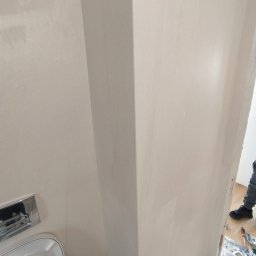 Natalii kuzyna - Świeżo wykończona ściana w łazience z widoczną spłuczką toaletową i fragmentem podłogi z narzędziami, ukazująca etap prac wykończeniowych.