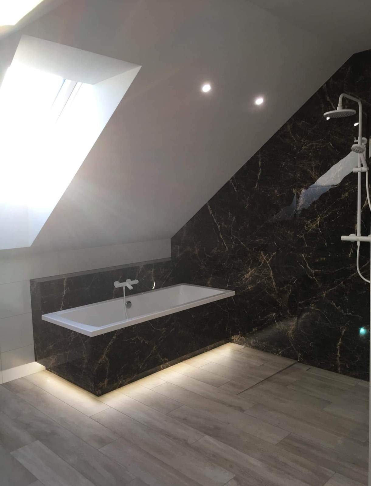 Nowoczesna łazienka na poddaszu z oknem dachowym, wykończona płytkami imitującymi czarny marmur z złotymi żyłami, podświetlana wanna i drewnopodobna podłoga.