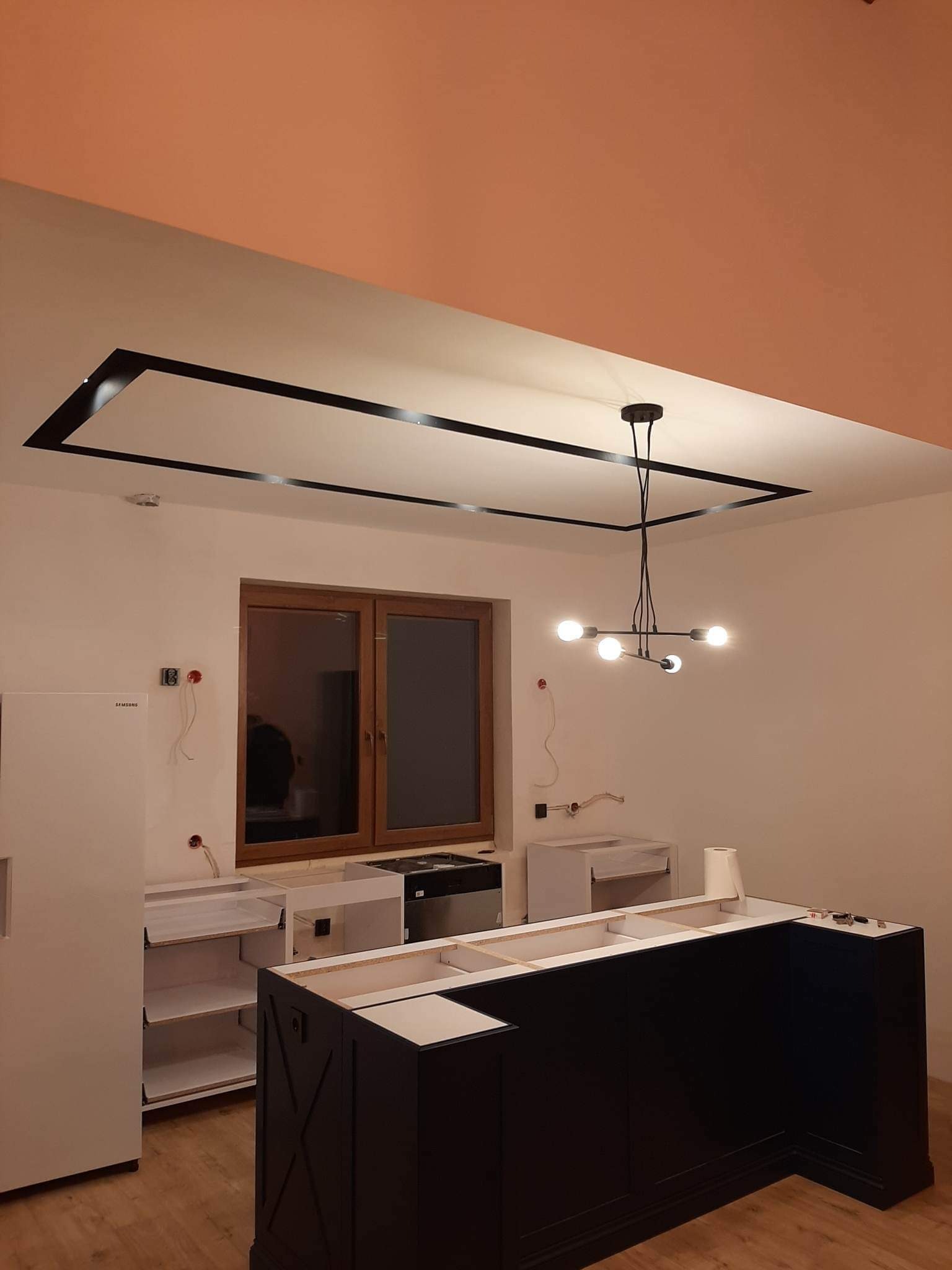 Wnętrze kuchni w trakcie montażu: czarne szafki wyspowe z białymi blatami, widoczne otwarte szafki, drewniane okno, nowoczesna lampa wisząca i czarna listwa dekoracyjna na suficie podwieszanym.