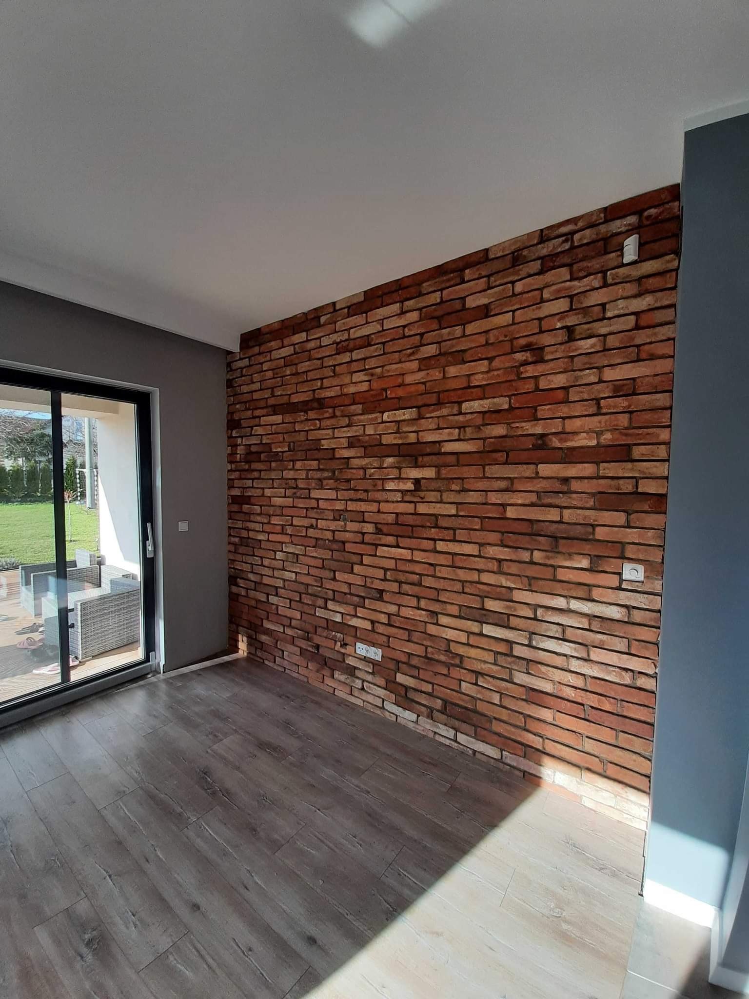 Wnętrze pokoju z dekoracyjną ścianą z czerwonej cegły, szarą ścianą, panelami podłogowymi w odcieniach szarości oraz przesuwnymi drzwiami balkonowymi z widokiem na taras i ogród.