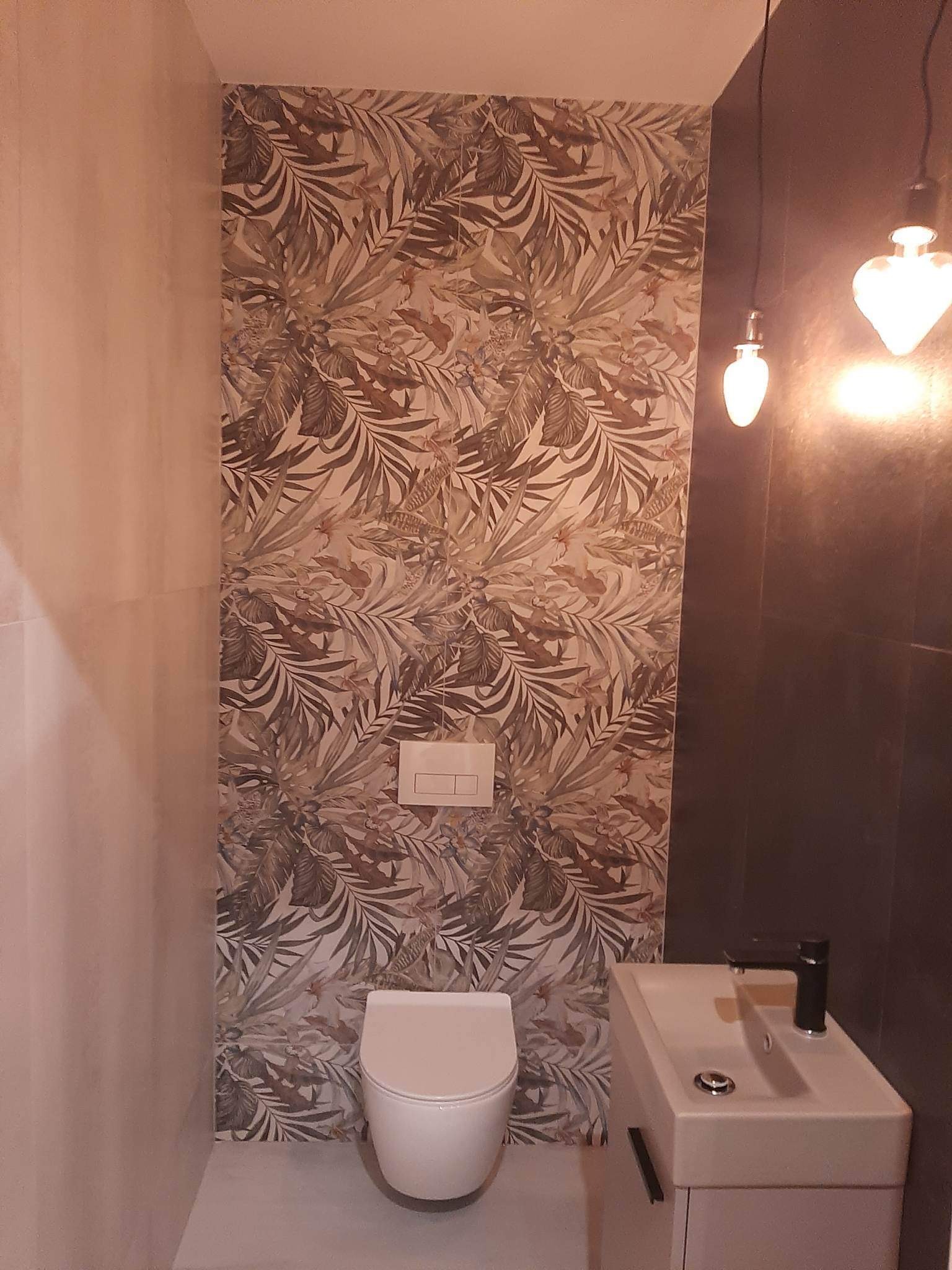 Nowoczesna toaleta z umywalką i baterią w kolorze czarnym, ściana wykończona płytkami z motywem liści, oświetlenie punktowe.