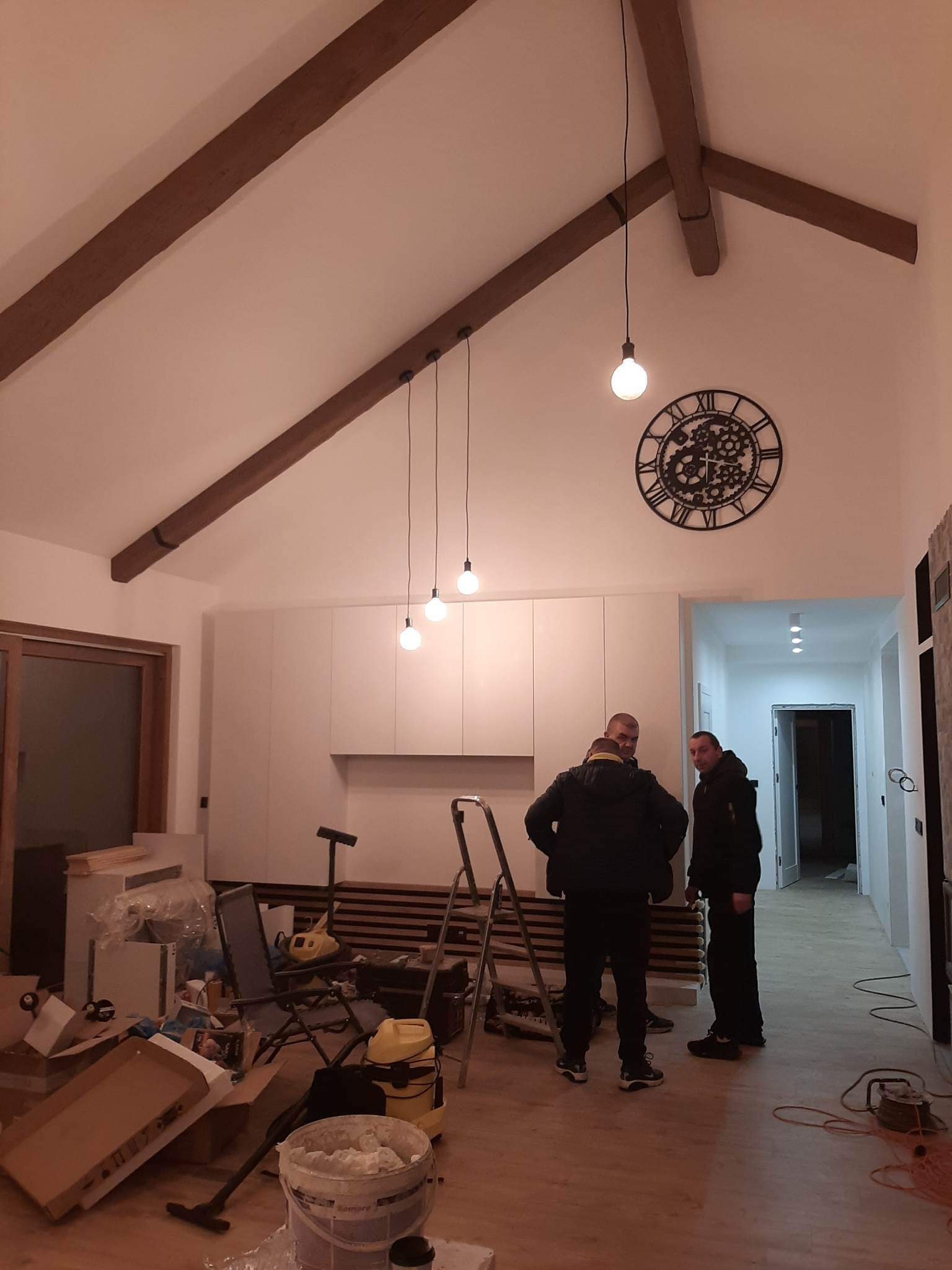 Wnętrze w trakcie prac wykończeniowych: drewniane belki stropowe, wiszące lampy, duży zegar na ścianie, widoczny bałagan i narzędzia, ekipa remontowa w tle.