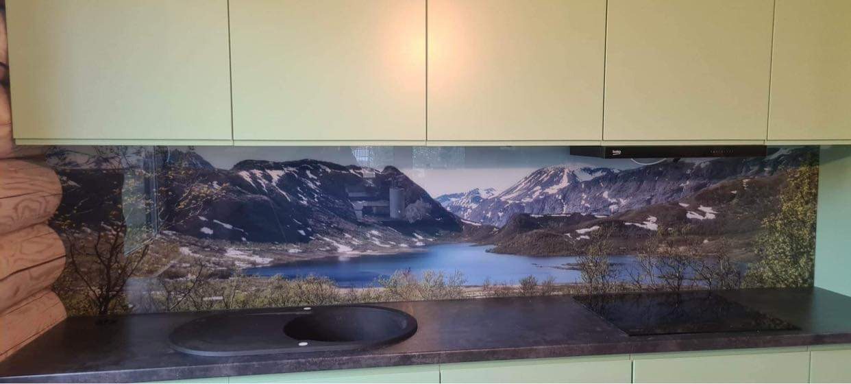 Szklana ściana nad blatem kuchennym z motywem górskiego krajobrazu z jeziorem, zamontowana między jasnymi szafkami a blatem z czarnym zlewem i płytą grzewczą.