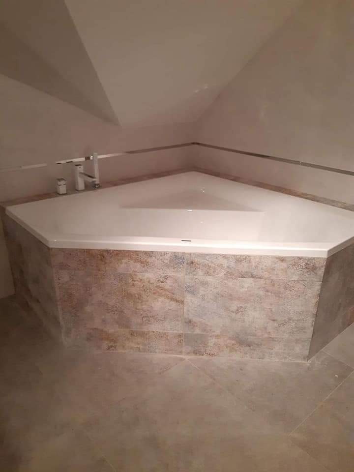 Trójkątna wanna obudowana płytkami w odcieniach beżu i brązu, z chromowaną baterią w narożnej łazience na poddaszu.