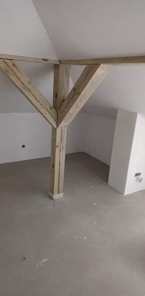 Surowe drewniane belki stropowe w nowym, surowym wnętrzu poddasza ze ścianami pomalowanymi na biało i szarą betonową podłogą.