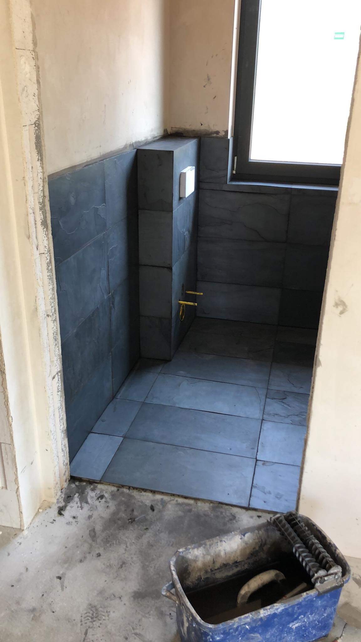 Wnętrze łazienki w trakcie wykańczania, z położonymi ciemnoszarymi płytkami na podłodze i ścianach, widoczna instalacja wodna i wiadro z narzędziami na pierwszym planie.
