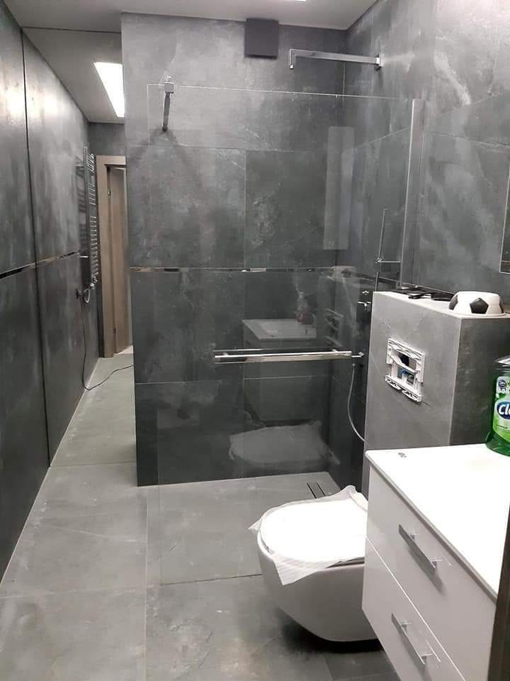 Nowoczesna łazienka z szarymi płytkami imitującymi kamień, kabina prysznicowa typu walk-in z przezroczystego szkła, podwieszana toaleta z papierem ochronnym na desce i biała szafka z szufladami.