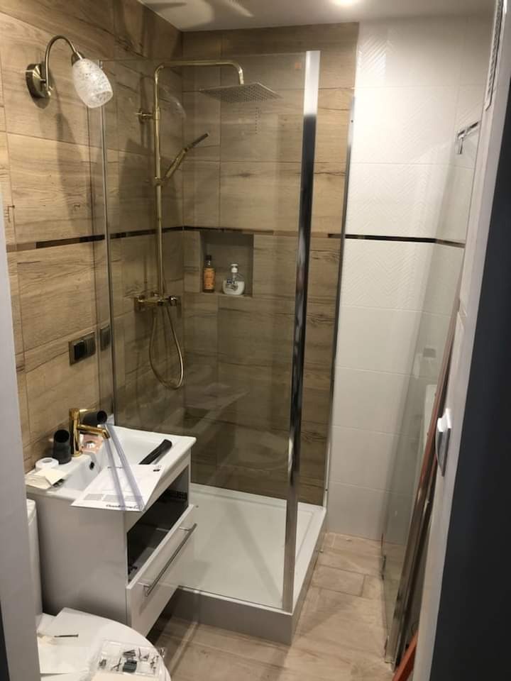 Nowoczesna kabina prysznicowa z złotą armaturą i odpływem liniowym, umywalka z szafką w trakcie montażu, widoczne narzędzia i instrukcja.