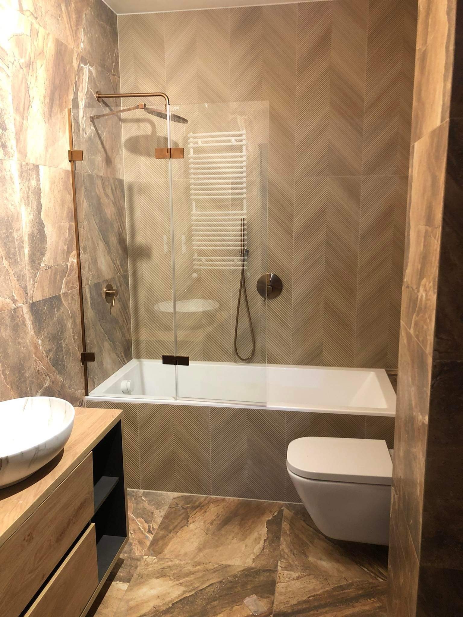 Wykończona łazienka z brązowymi płytkami imitującymi marmur i drewno, szklany parawan nawannowy z miedzianymi okuciami, wisząca toaleta.