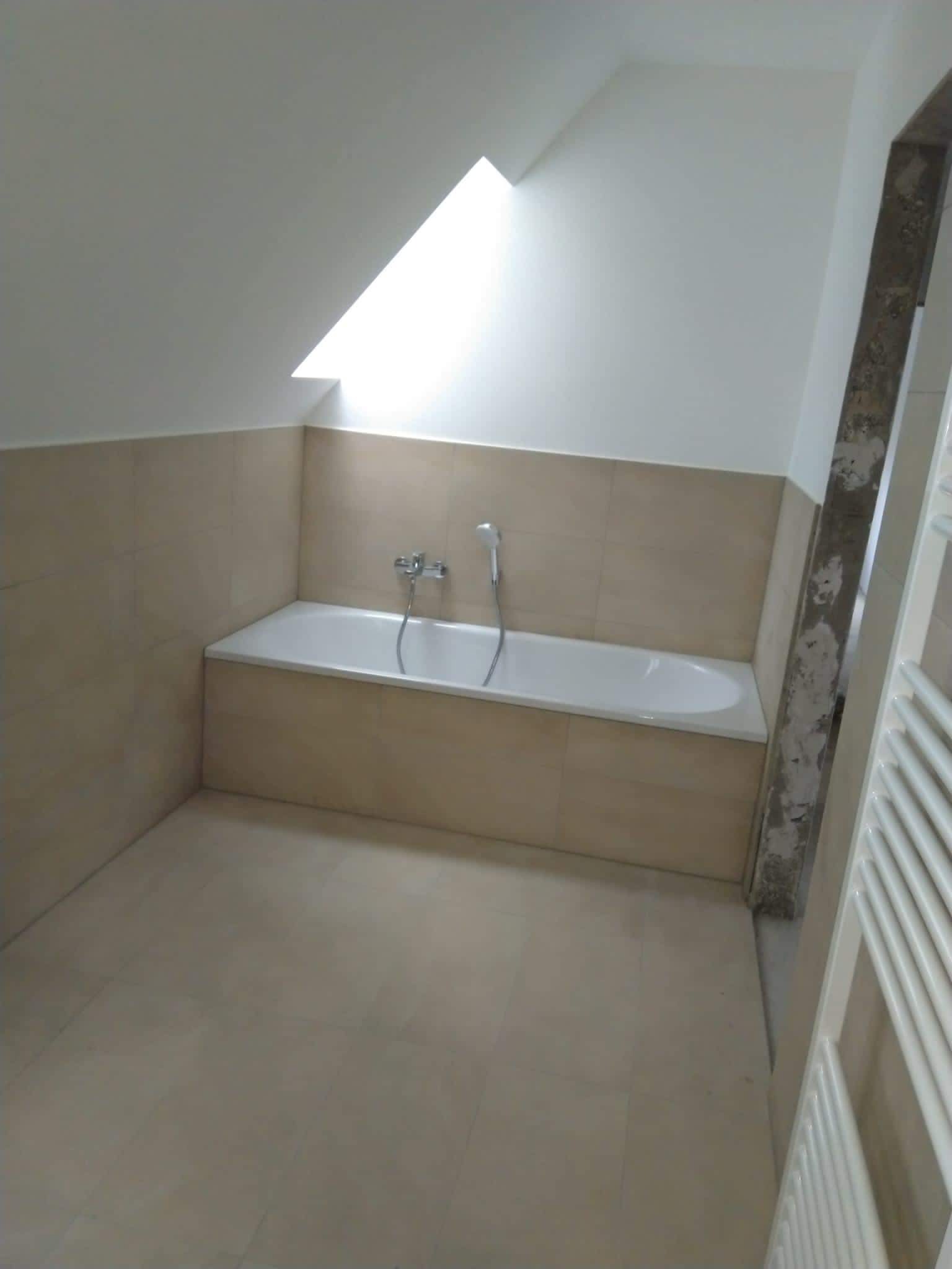 Łazienka w trakcie remontu, widoczna wanna obudowana płytkami w odcieniach beżu, fragment grzejnika drabinkowego i sufit ze skosem oraz okno dachowe, ściana przy drzwiach w trakcie wykończenia.