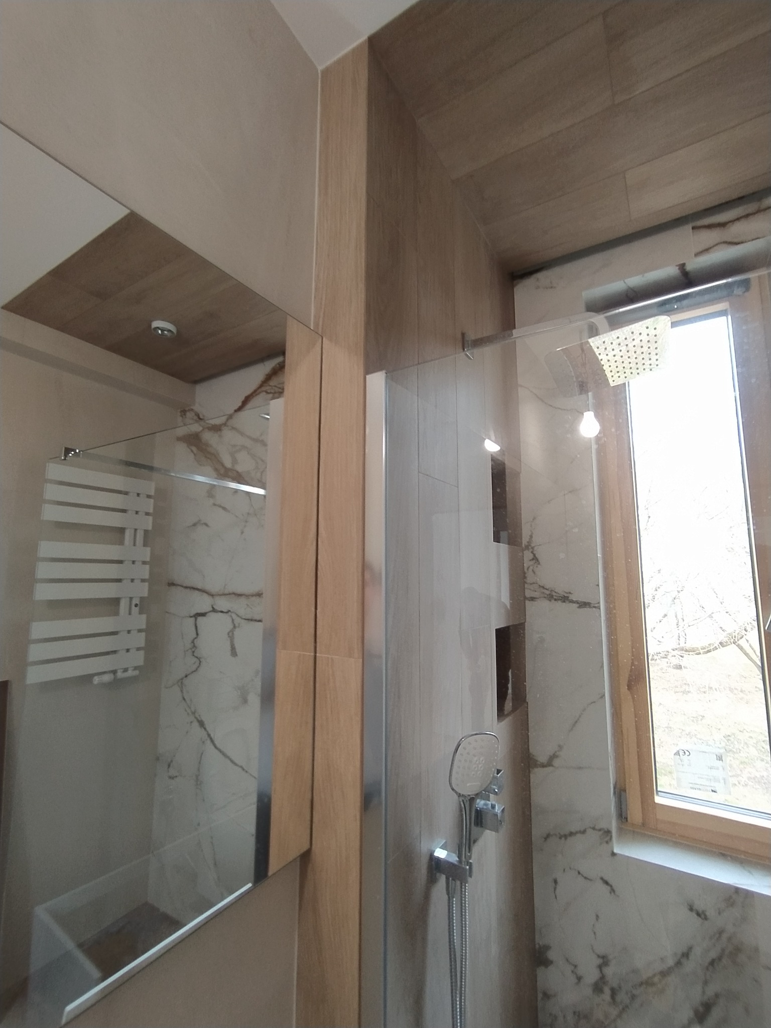 Szklana kabina prysznicowa typu walk-in z deszczownicą i słuchawką, marmurowe płytki na ścianach, drewnopodobna okładzina sufitu i ściany, grzejnik drabinkowy w odbiciu lustrzanym.