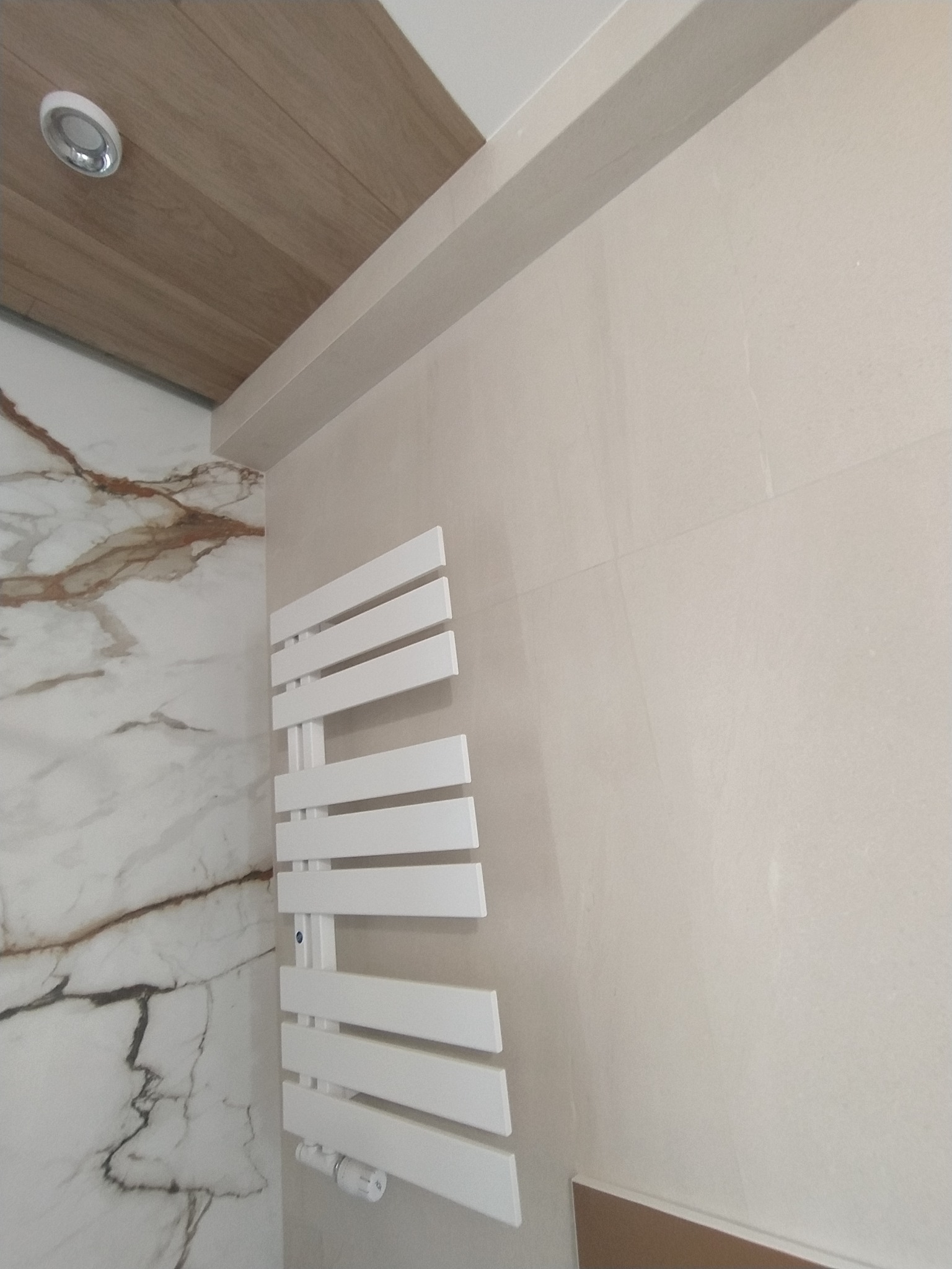 Biały grzejnik łazienkowy drabinkowy na tle jasnej, beżowej ściany z płytek, obok ściana z płytek imitujących marmur z brązowymi żyłami, widoczny fragment drewnianego sufitu z oświetleniem punktowym.