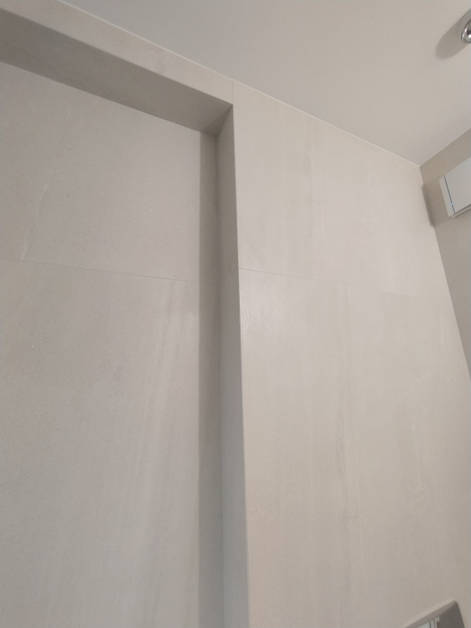 Ściana wyłożona jasnymi, beżowymi płytkami ceramicznymi z subtelną, poziomą fugą, widoczny fragment sufitu i klimatyzacji.