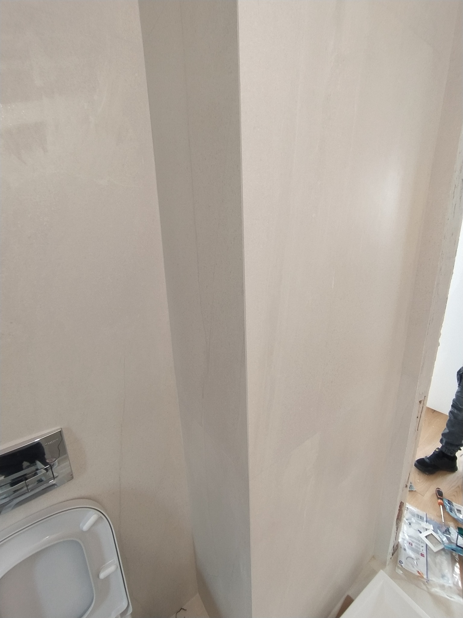 Świeżo wykończona ściana w łazience z widoczną spłuczką toaletową i fragmentem podłogi z narzędziami, ukazująca etap prac wykończeniowych.