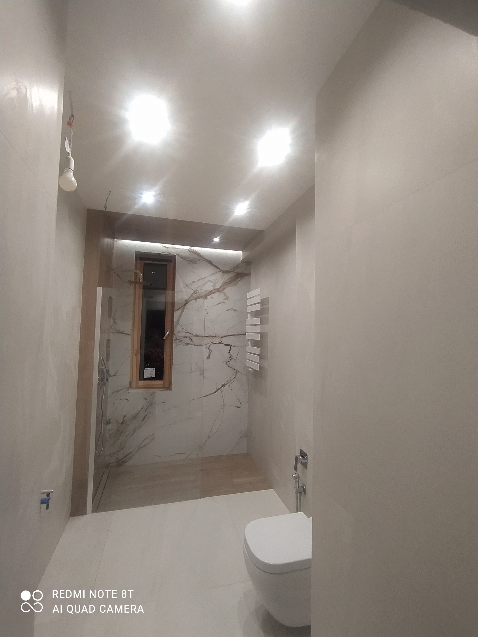 Nowoczesna łazienka z kabiną prysznicową typu walk-in z marmurowymi płytkami, wiszącą toaletą i grzejnikiem drabinkowym, oświetlona punktowymi lampami sufitowymi.