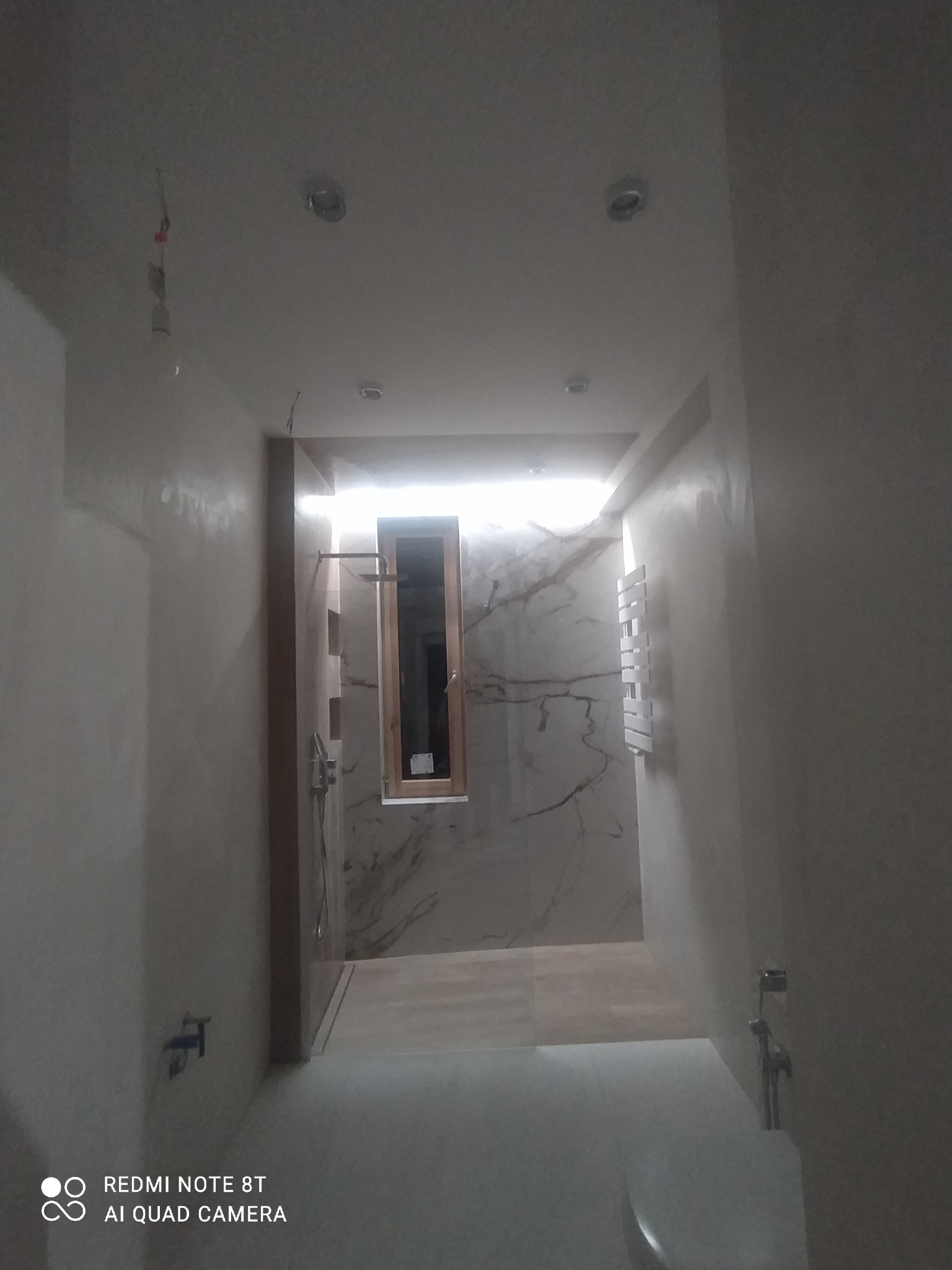 Wnętrze łazienki w trakcie remontu, widoczne surowe ściany przygotowane do malowania, fragment umywalki, grzejnik drabinkowy na ścianie wyłożonej płytkami imitującymi marmur, okno z drewnianą ramą...