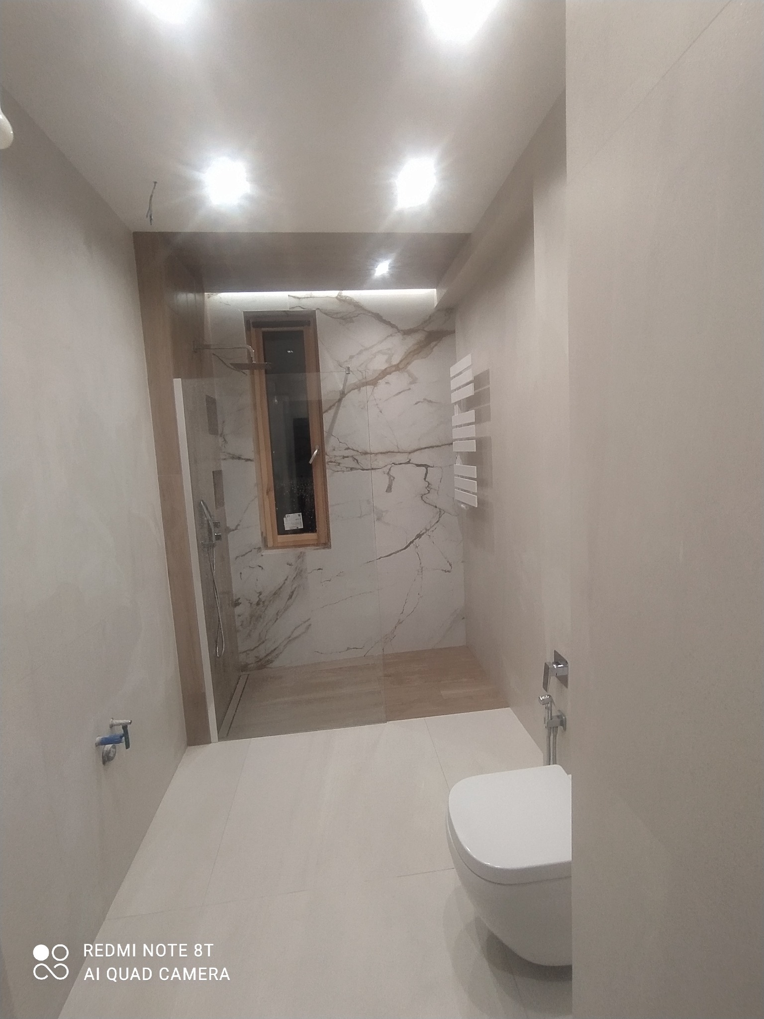 Nowoczesna łazienka z kabiną prysznicową typu walk-in, wykończona płytkami imitującymi marmur z brązowymi żyłami, podświetlane lustro, wisząca toaleta i grzejnik drabinkowy w kolorze białym.