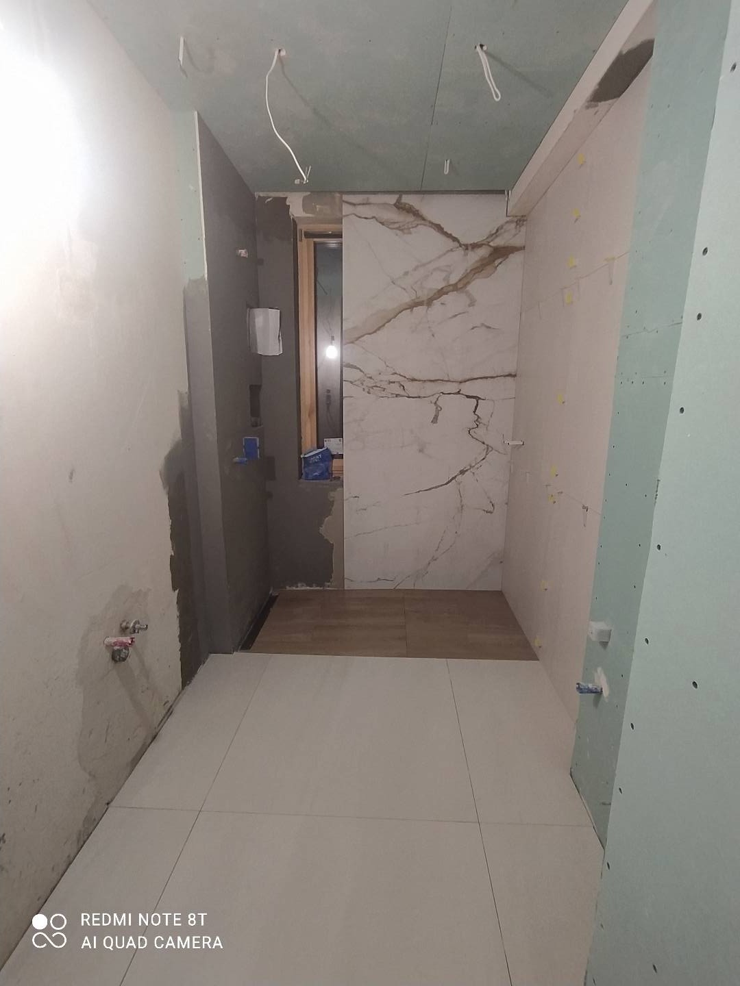 Łazienka w trakcie remontu: Częściowo położone białe płytki podłogowe, marmurowe płytki ścienne z brązowymi żyłami na ścianie prysznica, surowa ściana z instalacją hydrauliczną, odkryte kable...