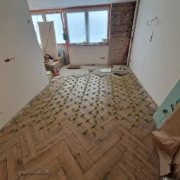 Układanie drewnopodobnych płytek podłogowych w jodełkę, z użyciem zielonych krzyżyków dystansowych, na tle surowej ściany z cegły i okien zabezpieczonych folią.