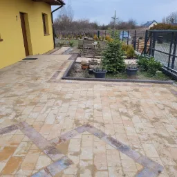 Nowo ułożona, piaskowana kostka brukowa w odcieniach beżu i brązu z dekoracyjnym wzorem, otoczona obrzeżem z ciemnej kostki, przylegająca do żółtej elewacji domu z numerem 7 i fragmentem ogrodu...