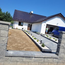 MG Mirosław Geisler - Nowoczesny podjazd z kostki brukowej prowadzący do domu z garażem, widoczny mężczyzna i niebieski bus, fragment murku z kostki po lewej stronie, część terenu przygotowana pod nasadzenia.