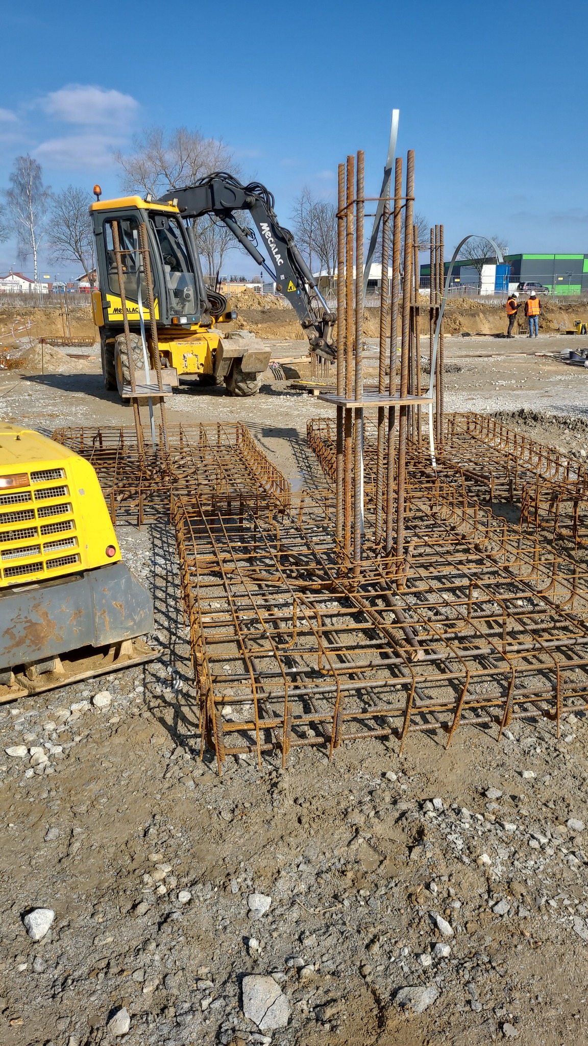 Żółta koparka Mecalac przygotowuje teren pod fundament zbrojony stalowymi prętami na budowie w słoneczny dzień.