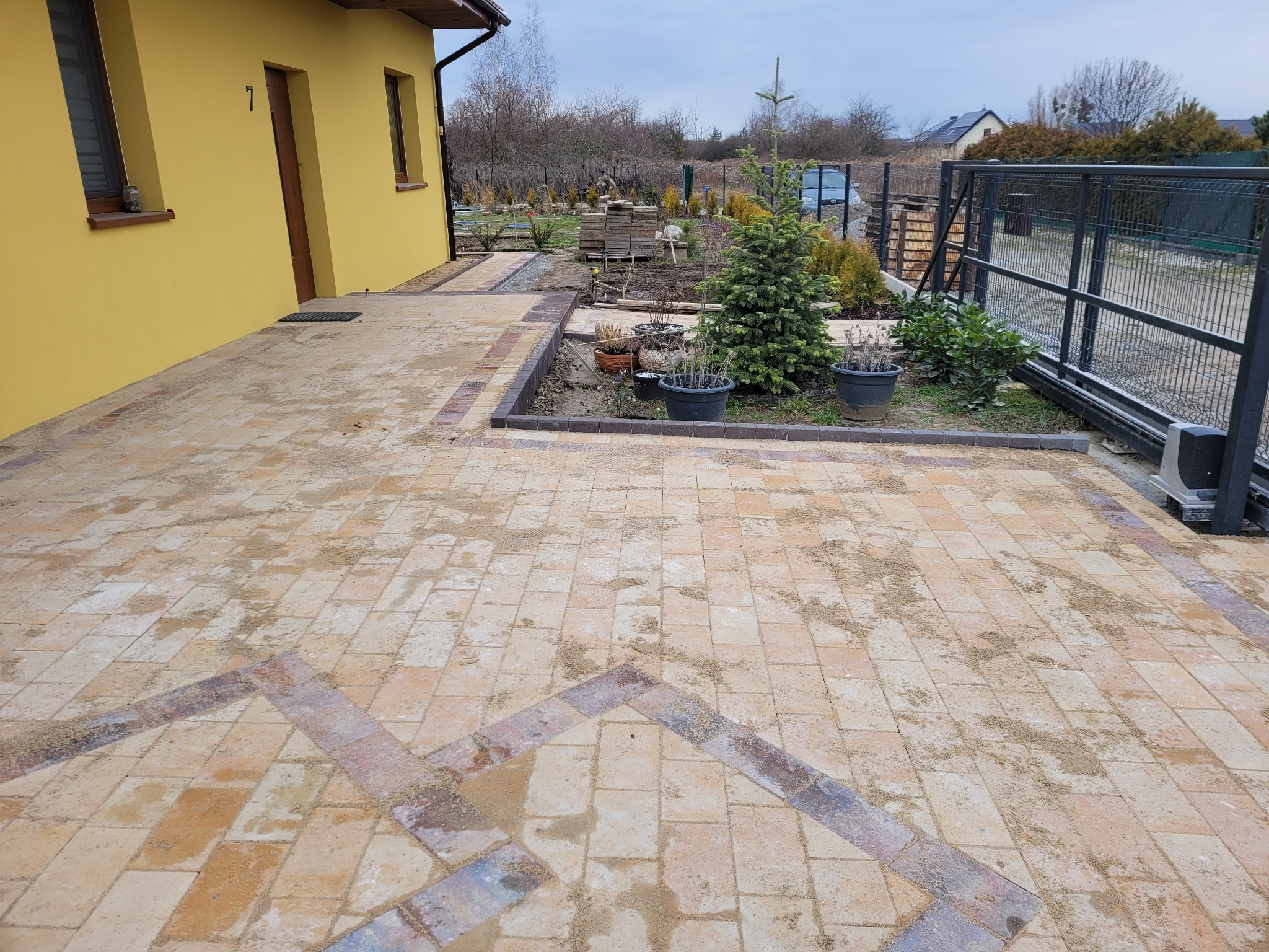 Nowo ułożona, piaskowana kostka brukowa w odcieniach beżu i brązu z dekoracyjnym wzorem, otoczona obrzeżem z ciemnej kostki, przylegająca do żółtej elewacji domu z numerem 7 i fragmentem ogrodu...