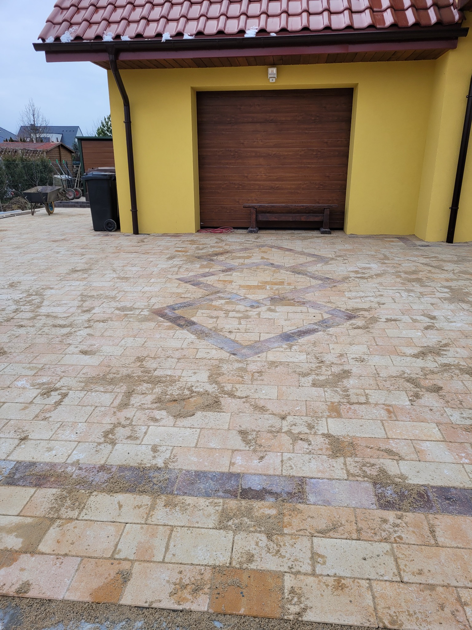 Ułożona kostka brukowa w odcieniach beżu i brązu z dekoracyjnym wzorem rombów, częściowo zasypana piaskiem, na tle żółtego garażu z drewnianą bramą i ławką.