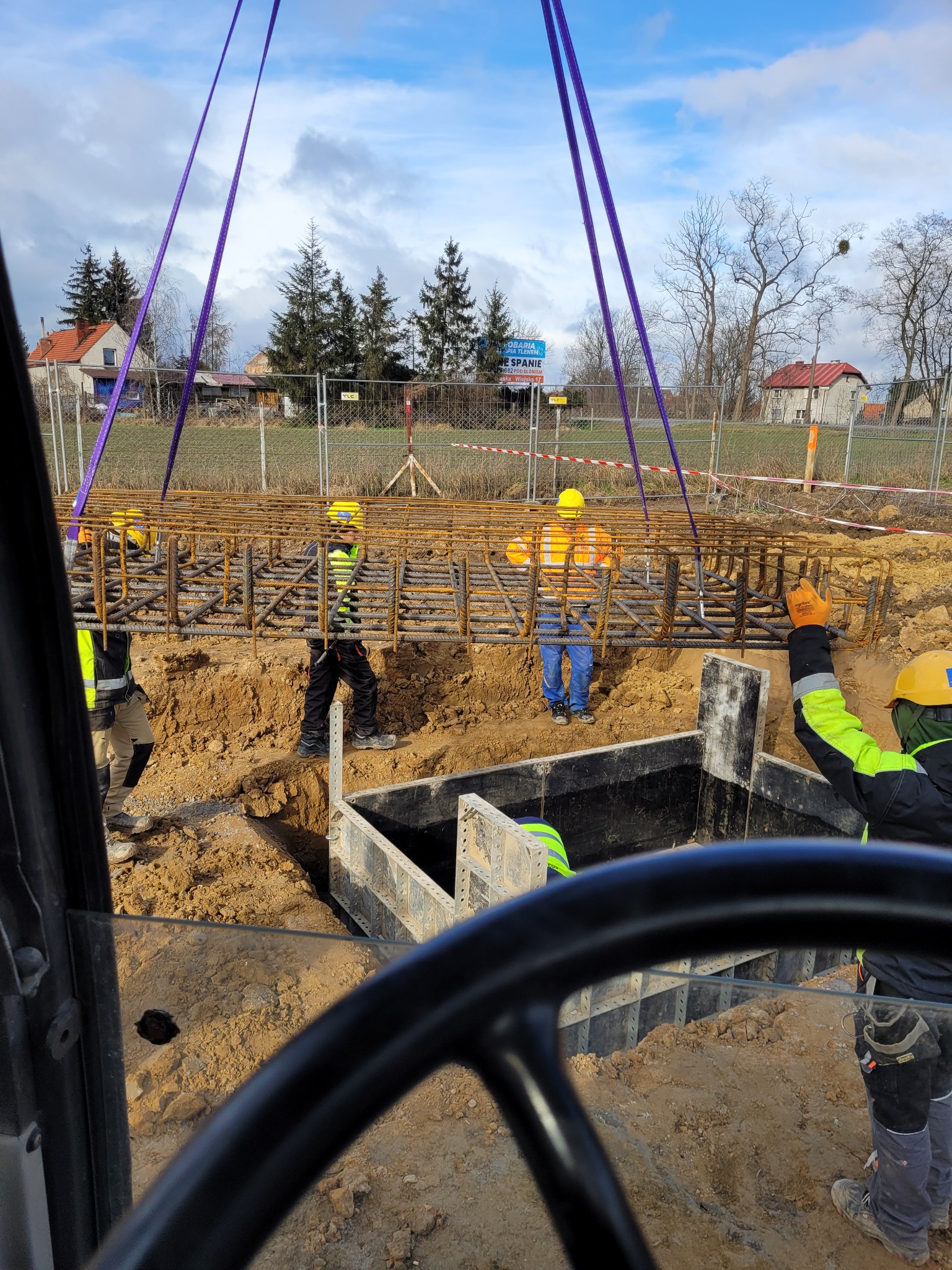 Pracownicy w kaskach i kamizelkach odblaskowych za pomocą pasów transportowych opuszczają zbrojenie do wykopu z szalunkiem, widok z kabiny operatora maszyny.