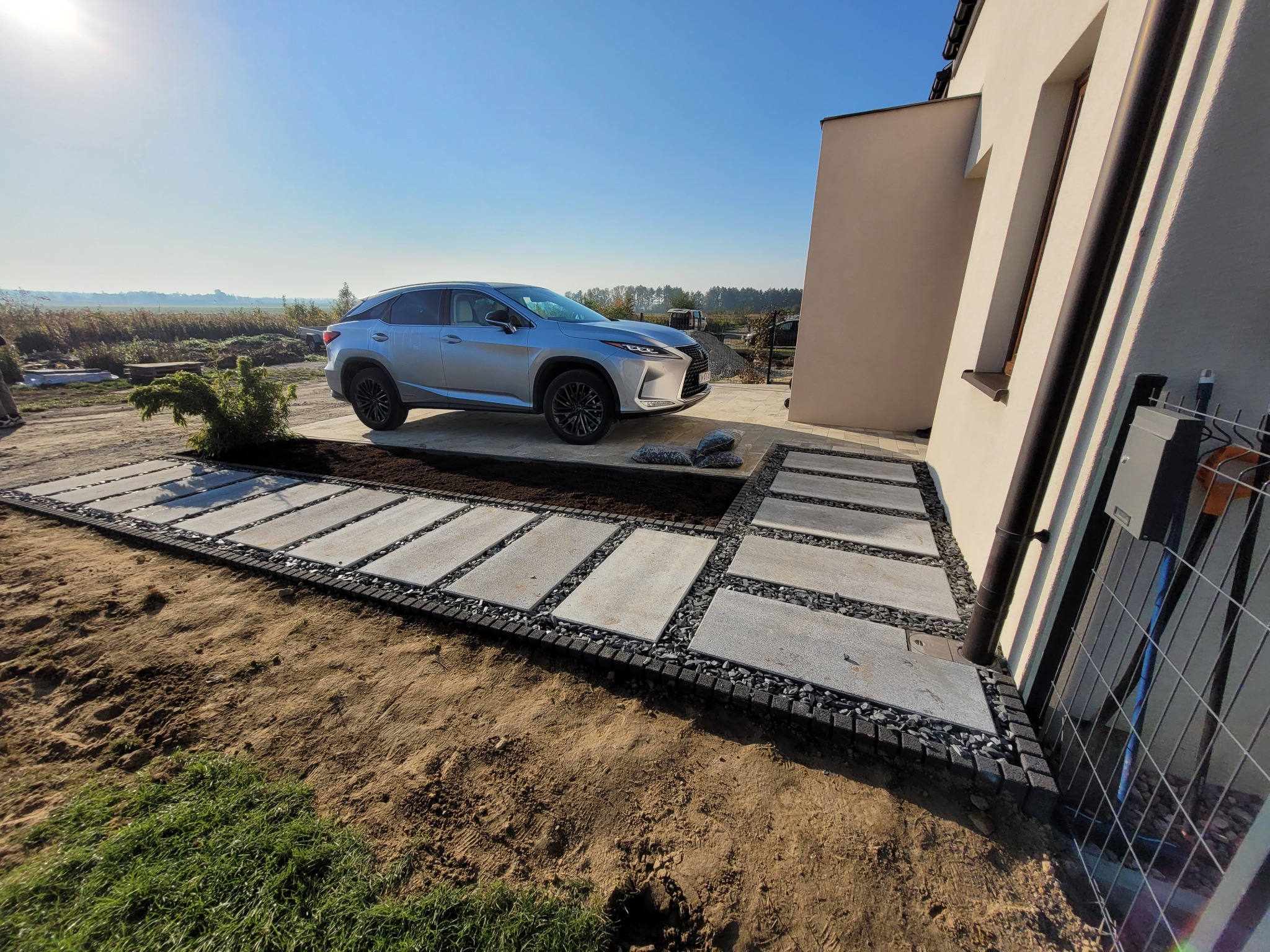 Nowoczesna ścieżka z szarych płyt betonowych oddzielonych czarnym grysem, prowadząca do domu z zaparkowanym srebrnym SUV-em, widok w słoneczny dzień.