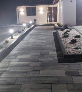 Oświetlona ścieżka z szarej kostki brukowej prowadząca do wejścia domu, z rabatami po bokach wypełnionymi białym żwirem i niskimi roślinami, nocne ujęcie.