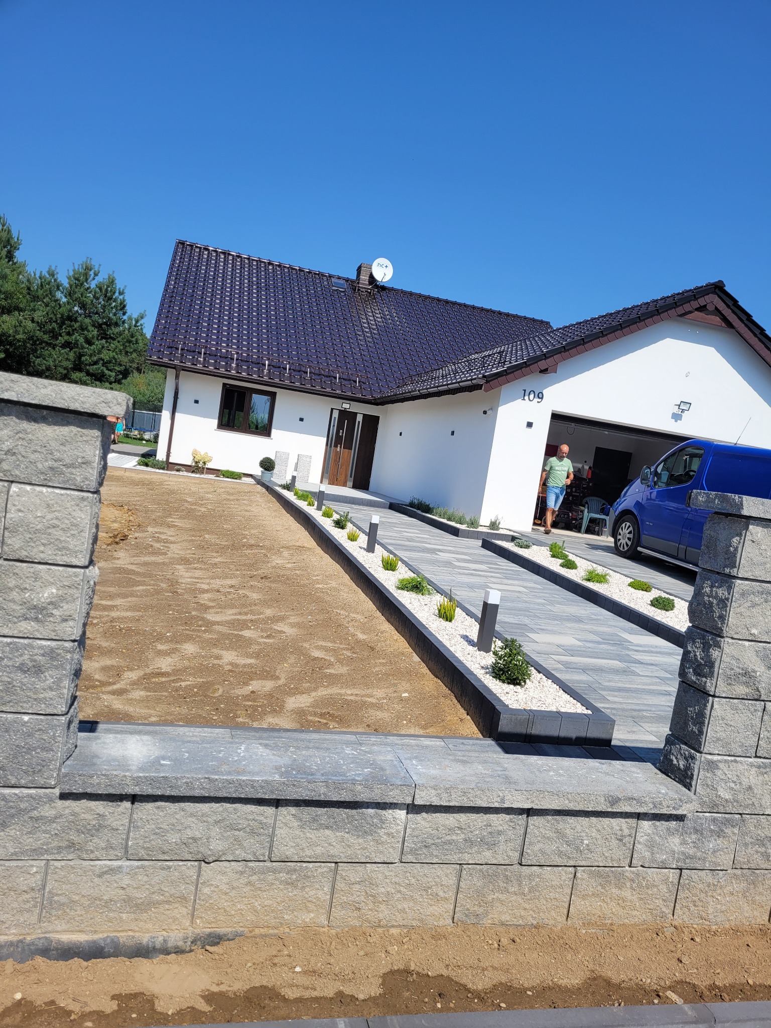 Nowoczesny podjazd z kostki brukowej prowadzący do domu z garażem, widoczny mężczyzna i niebieski bus, fragment murku z kostki po lewej stronie, część terenu przygotowana pod nasadzenia.