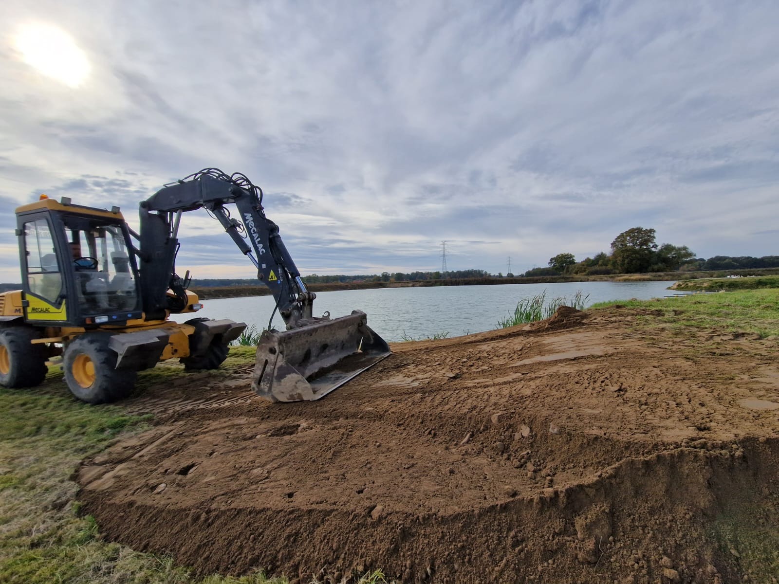 Żółto-czarna koparka kołowa Mecalac przy brzegu stawu, świeżo wykopana ziemia, pochmurne niebo.