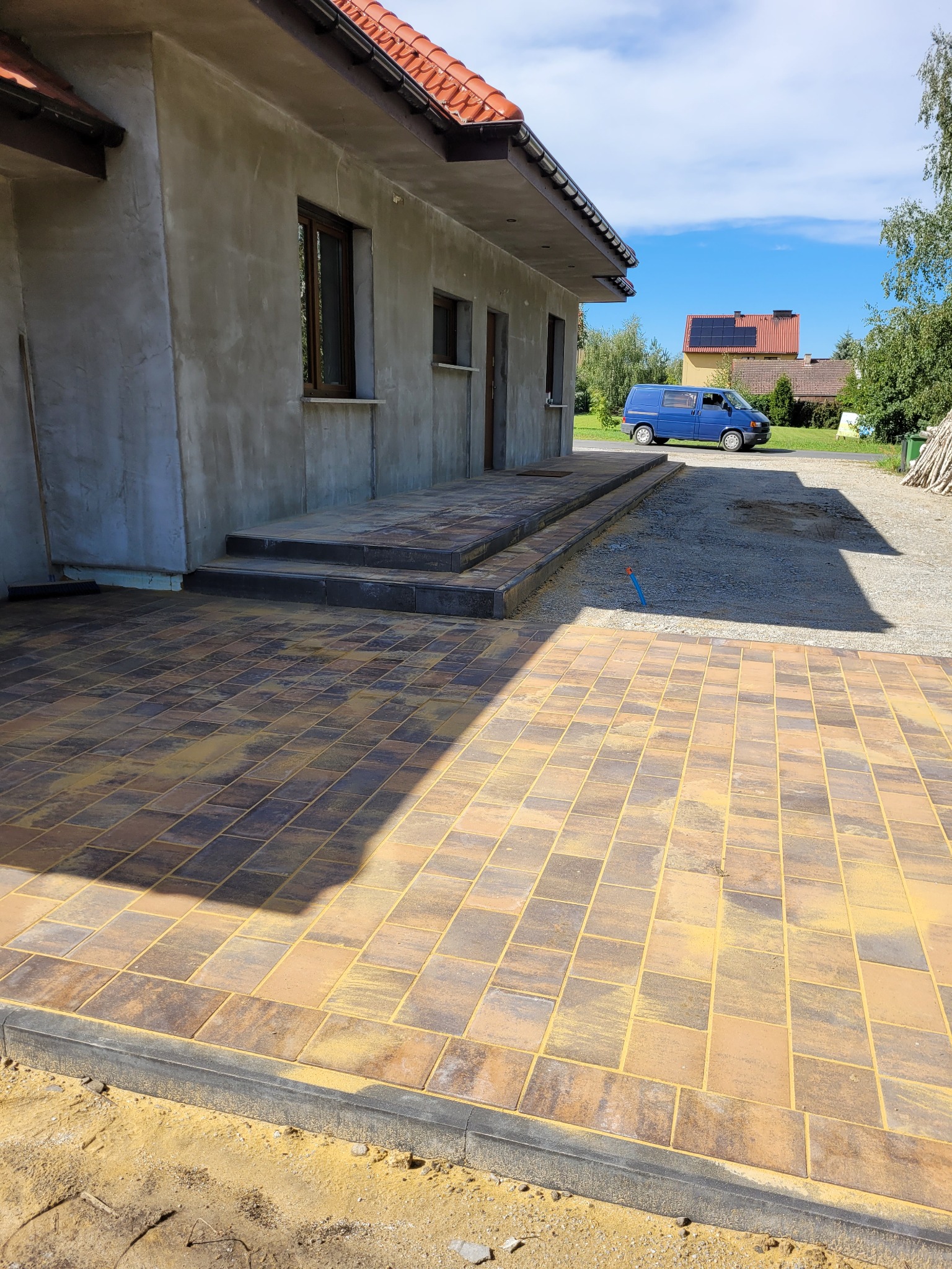 Ułożona kostka brukowa w odcieniach brązu i beżu przed budynkiem w stanie surowym, widoczne schody z kostki i niebieski bus w tle.