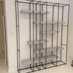 Farbud - Metalowa konstrukcja regału z asymetrycznie rozmieszczonymi szarymi półkami na tle białej ściany i podłogi z płytek.