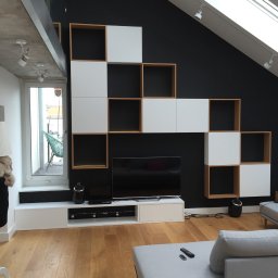 Farbud - Nowoczesny salon na poddaszu z czarną ścianą, drewnianą podłogą i biało-drewnianą zabudową meblową w kształcie schodów, podświetlany sufit z oknami dachowymi i widoczne elementy szarej sofy