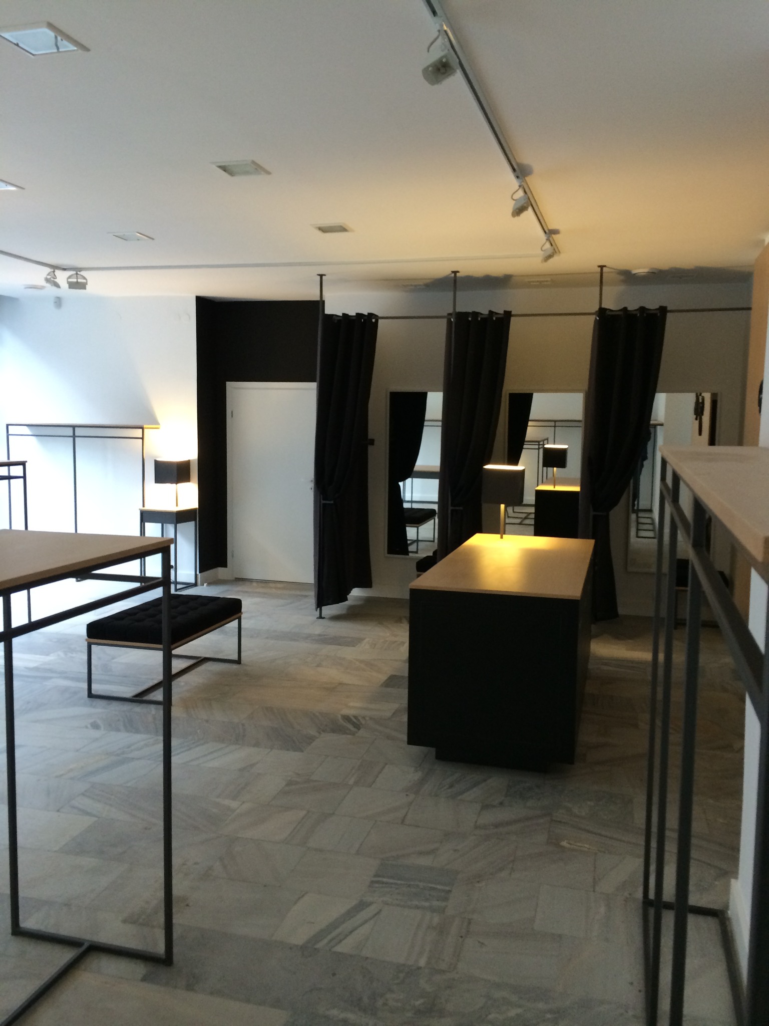 Wnętrze przymierzalni w sklepie odzieżowym z czarnymi zasłonami, marmurową podłogą, czarnymi meblami z drewnianymi blatami i lampami