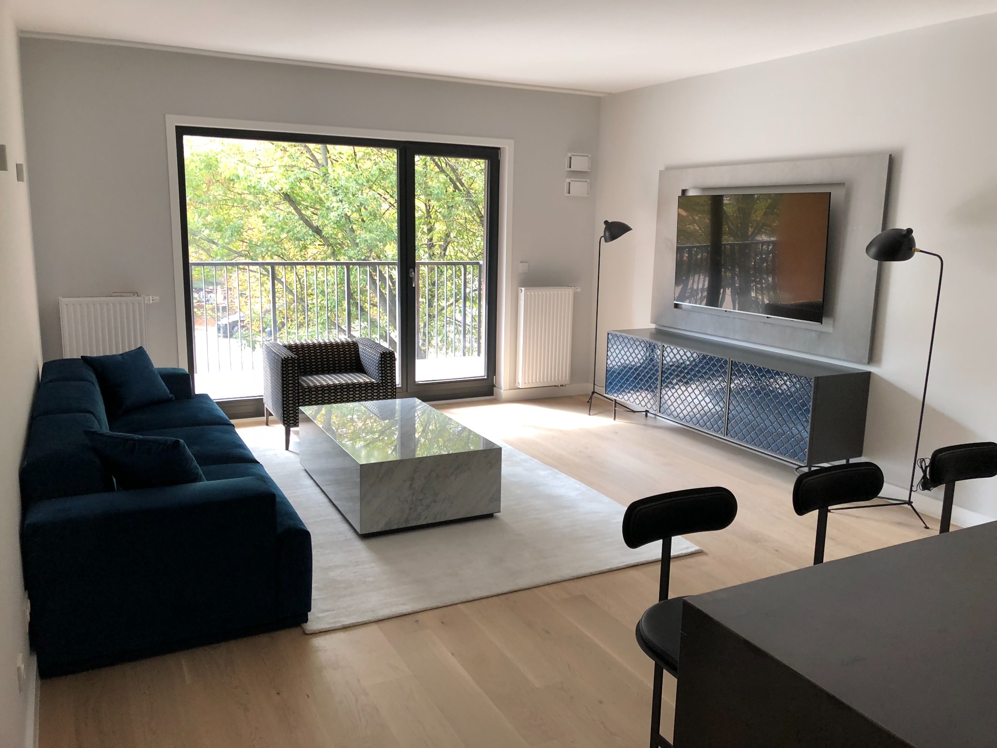 Nowoczesny salon z ciemnoniebieską sofą modułową, marmurowym stolikiem kawowym, szafką RTV z niebieskimi drzwiczkami i dużym oknem balkonowym z widokiem na drzewa.