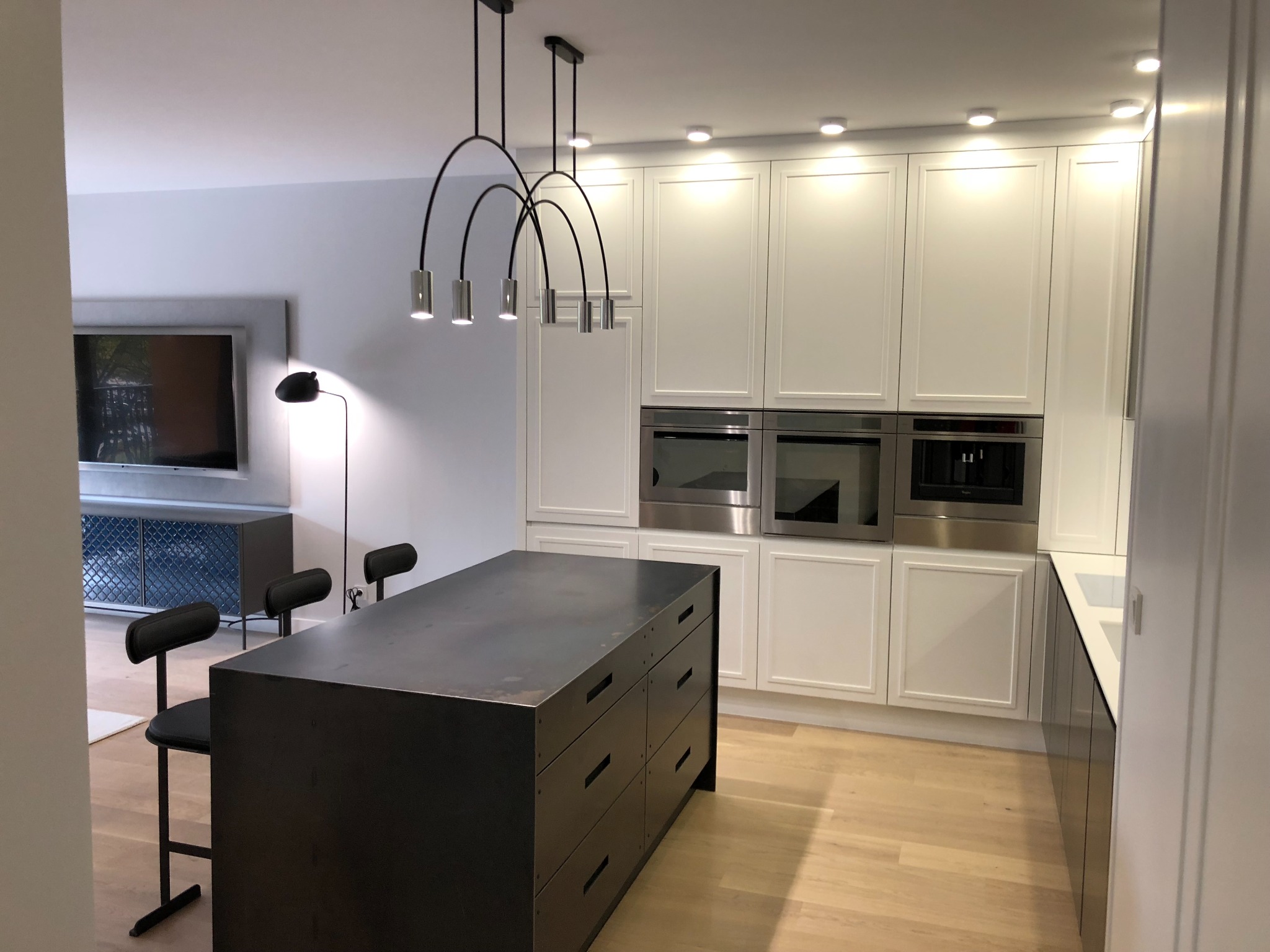 Nowoczesna kuchnia z wyspą, białymi szafkami z frezowanymi frontami i wbudowanymi sprzętami AGD, oświetlona designerskimi lampami łukowymi.