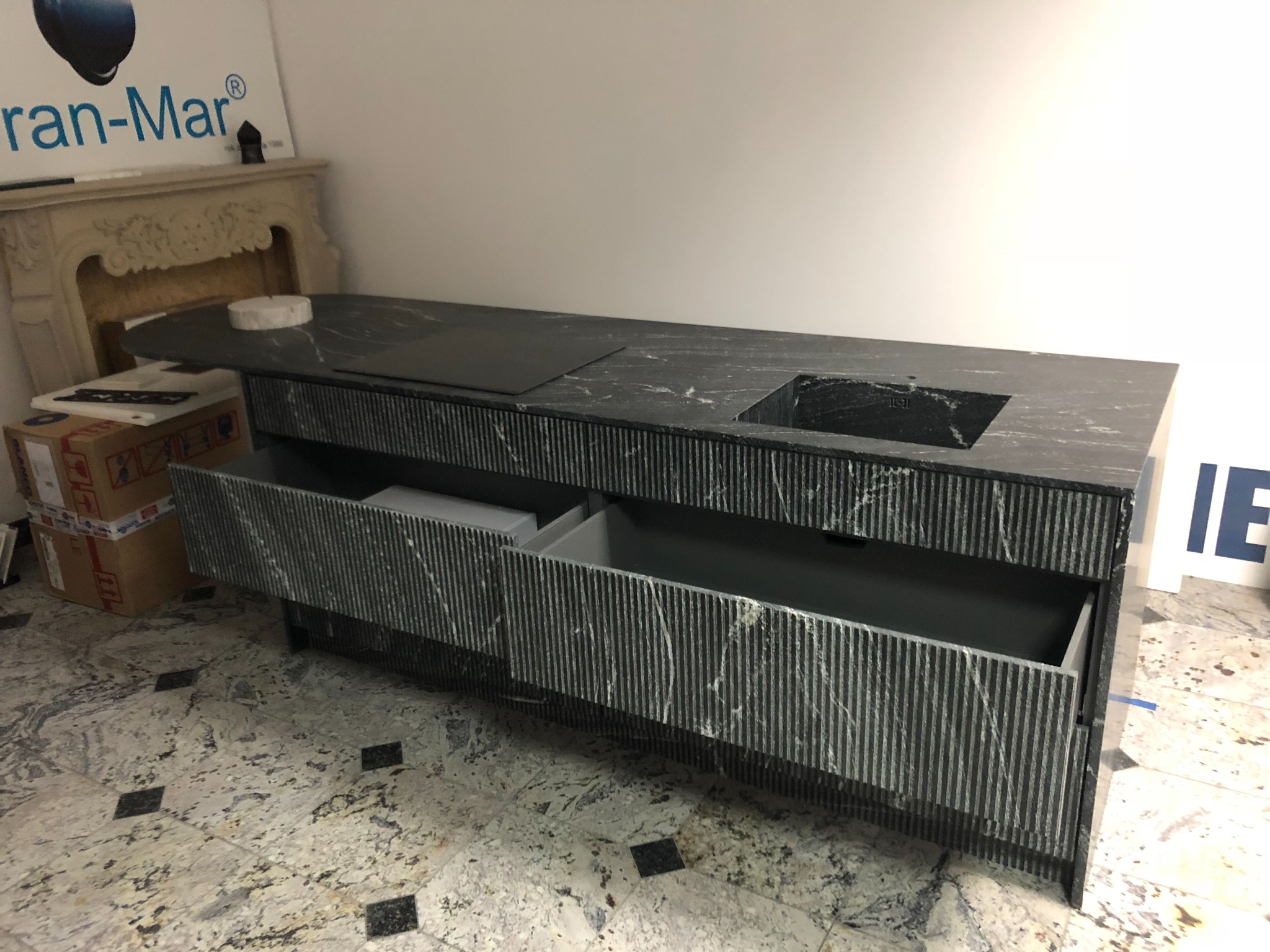 Długa szafka łazienkowa z marmurowym blatem, wbudowanym zlewem i płytą indukcyjną, z otwartymi szufladami i dekoracyjną fakturą pionowych pasków, na tle marmurowej podłogi i fragmentu reklamy.