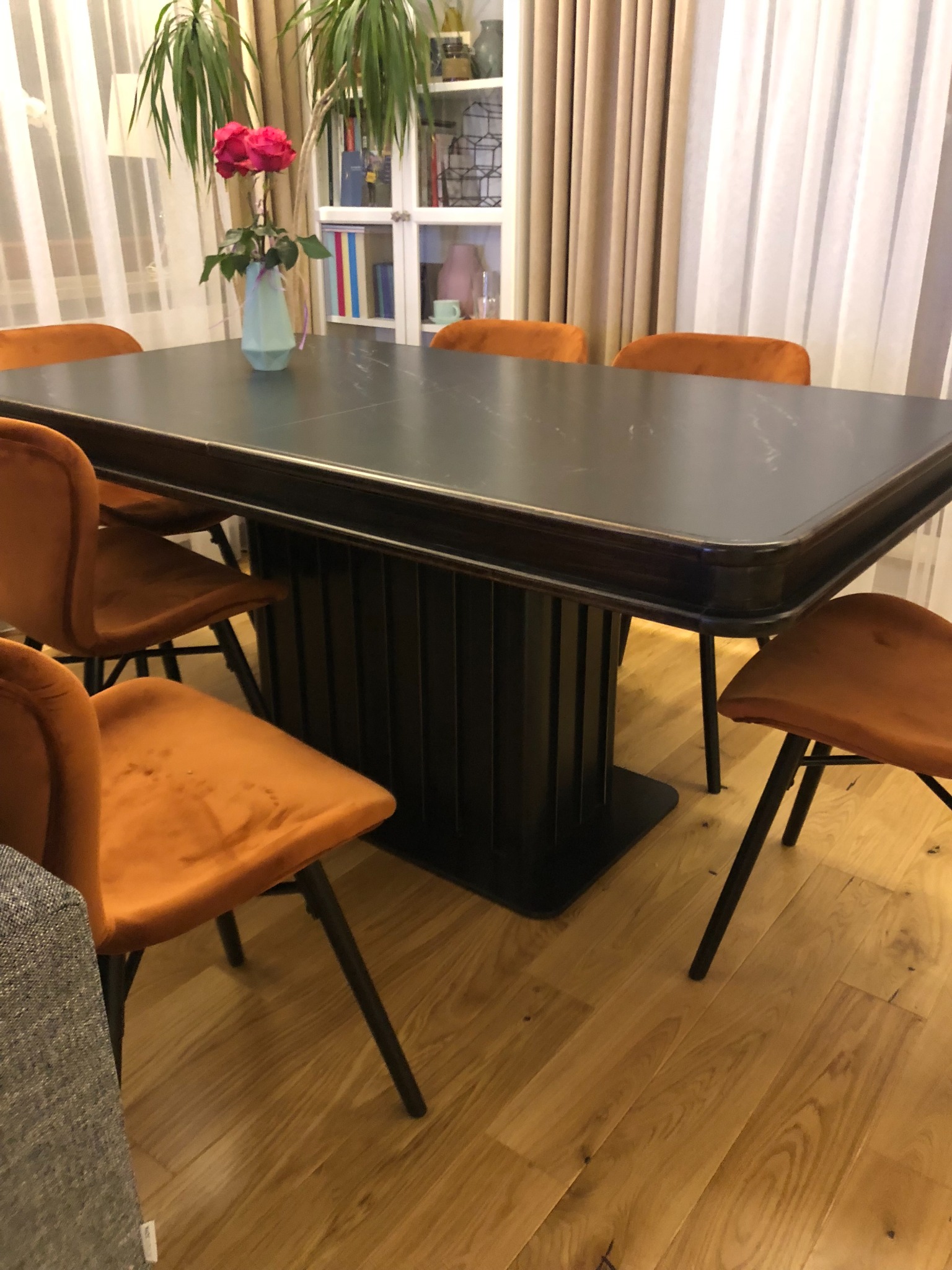 Stół z ciemnym blatem i podłużnymi żłobieniami w podstawie, otoczony pomarańczowymi krzesłami na cienkich, czarnych nogach. W tle widoczna roślina doniczkowa i biały regał z książkami.