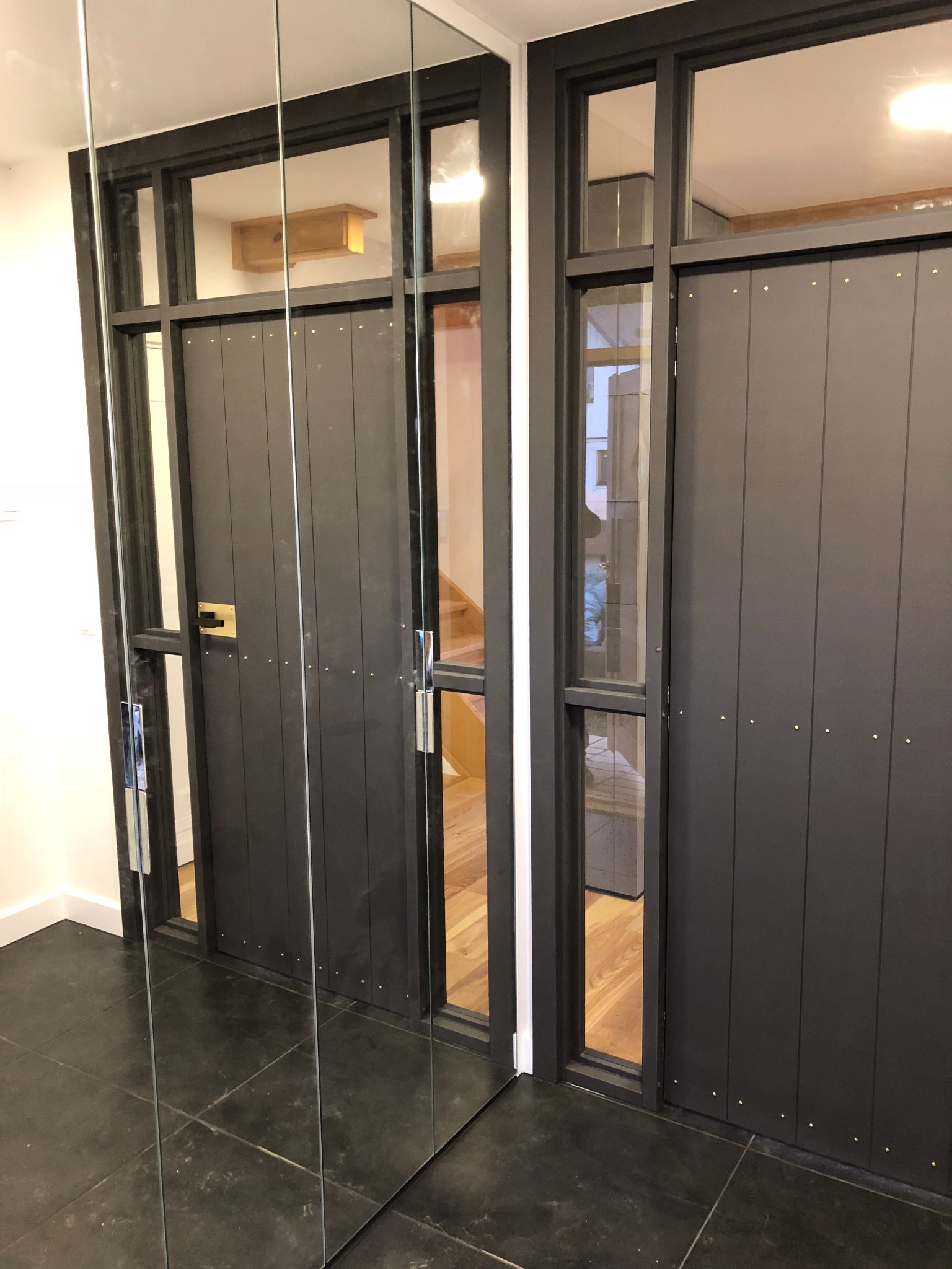 Nowoczesne, grafitowe drzwi z przeszkleniami i metalowymi detalami, widoczne na tle ciemnej podłogi i lustrzanej ściany.