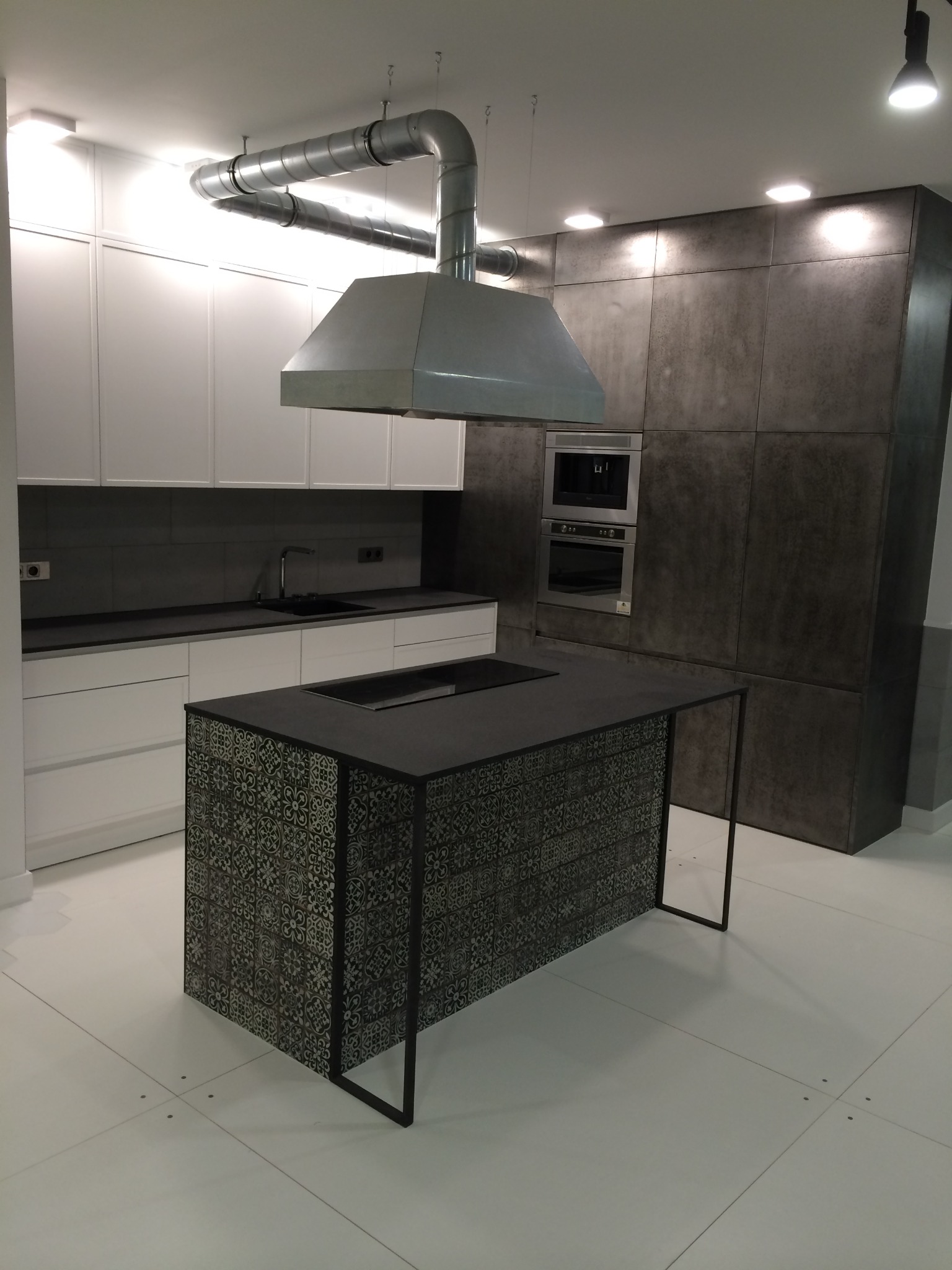 Nowoczesna kuchnia z wyspą wykończoną dekoracyjnymi płytkami w odcieniach szarości, białe szafki wiszące i szara zabudowa kuchenna z wbudowanym piekarnikiem i ekspresem do kawy.