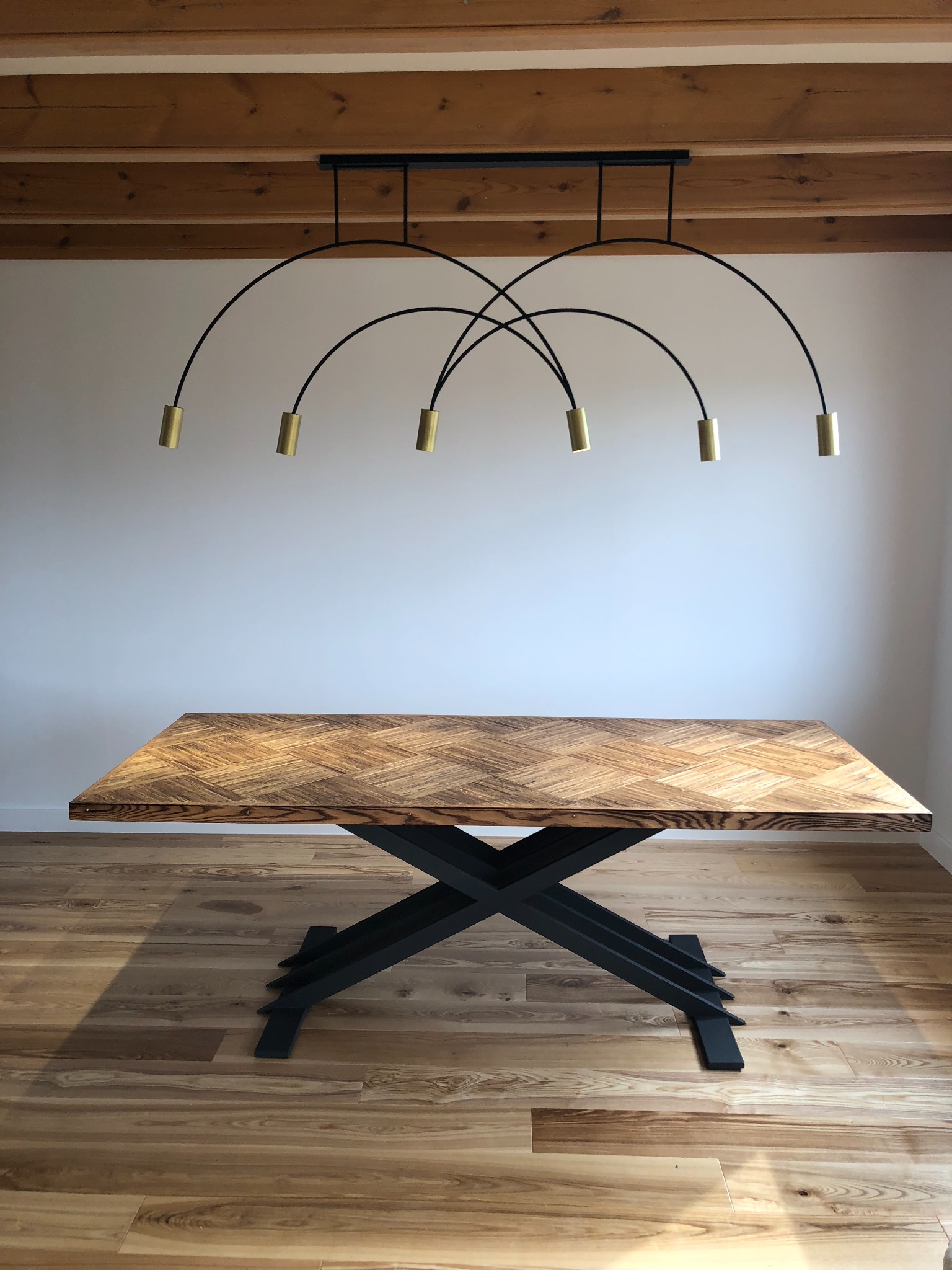 Stół z blatem z drewna ułożonego w jodełkę, na czarnej, metalowej podstawie w kształcie litery X, oświetlony nowoczesną lampą z sześcioma złotymi reflektorami na łukowatych ramionach, na tle białej...