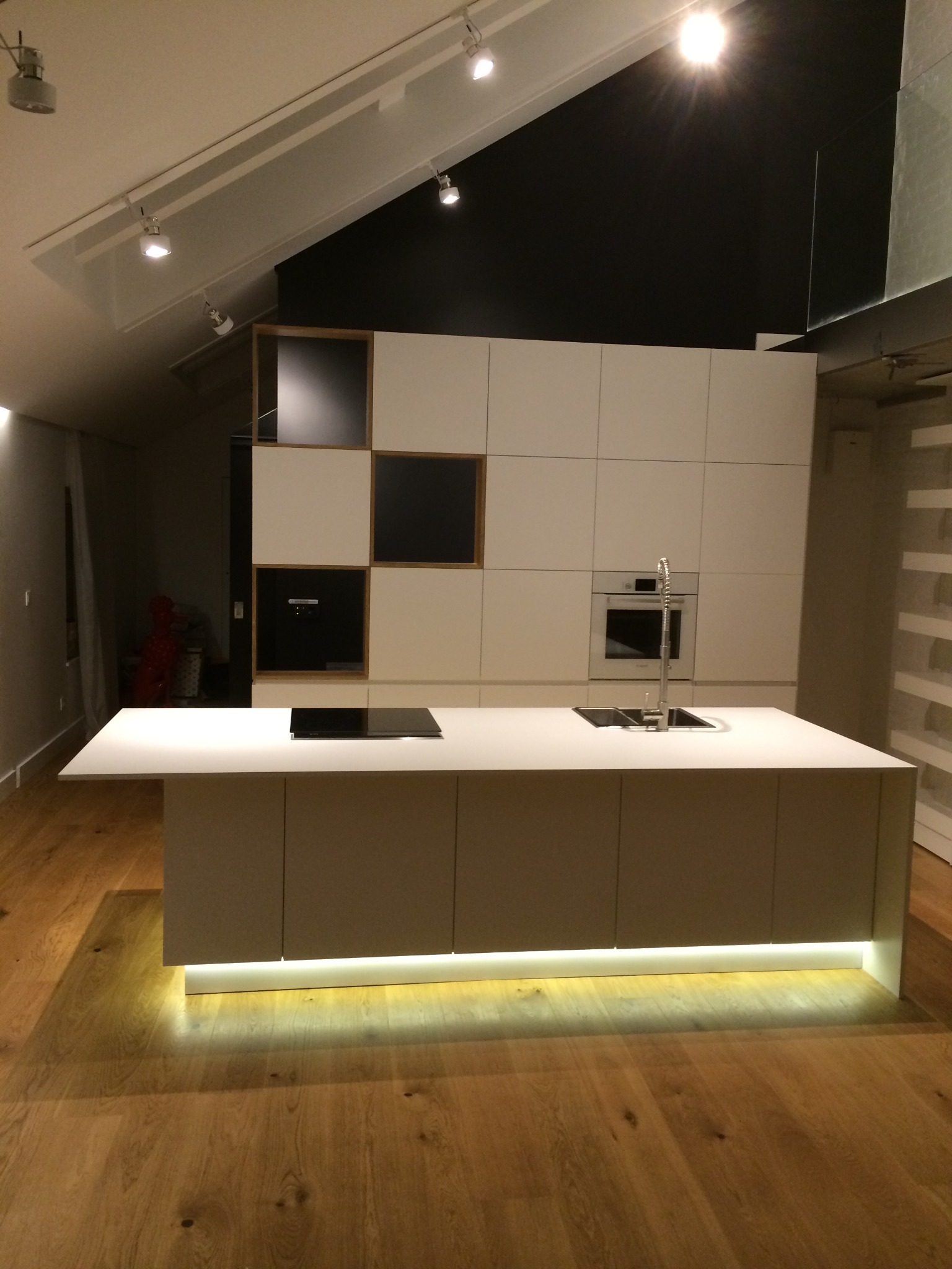 Nowoczesna kuchnia z wyspą oświetloną od dołu, białe szafki bez uchwytów, drewniana podłoga i czarna ściana w tle.