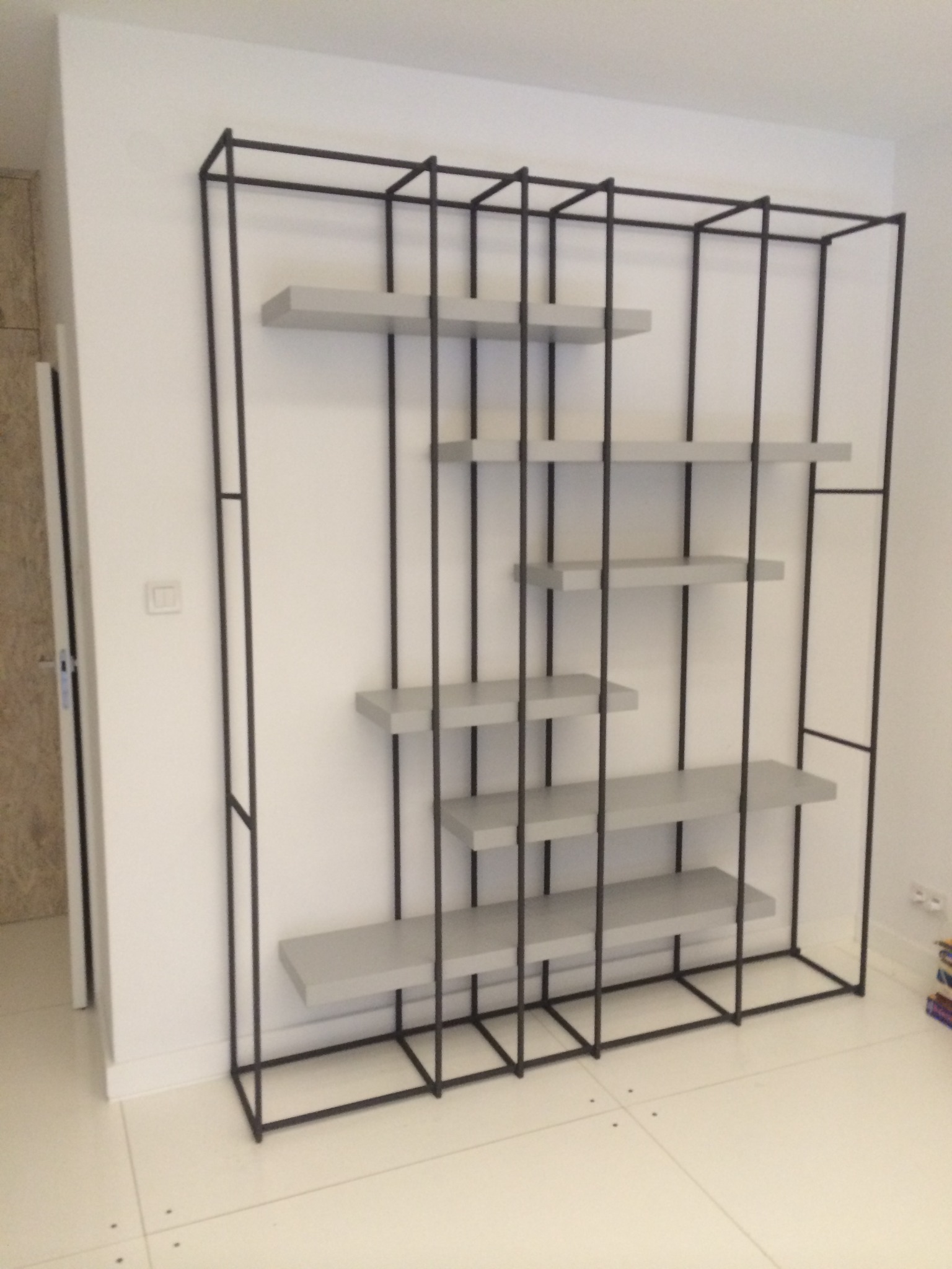 Metalowa konstrukcja regału z asymetrycznie rozmieszczonymi szarymi półkami na tle białej ściany i podłogi z płytek.