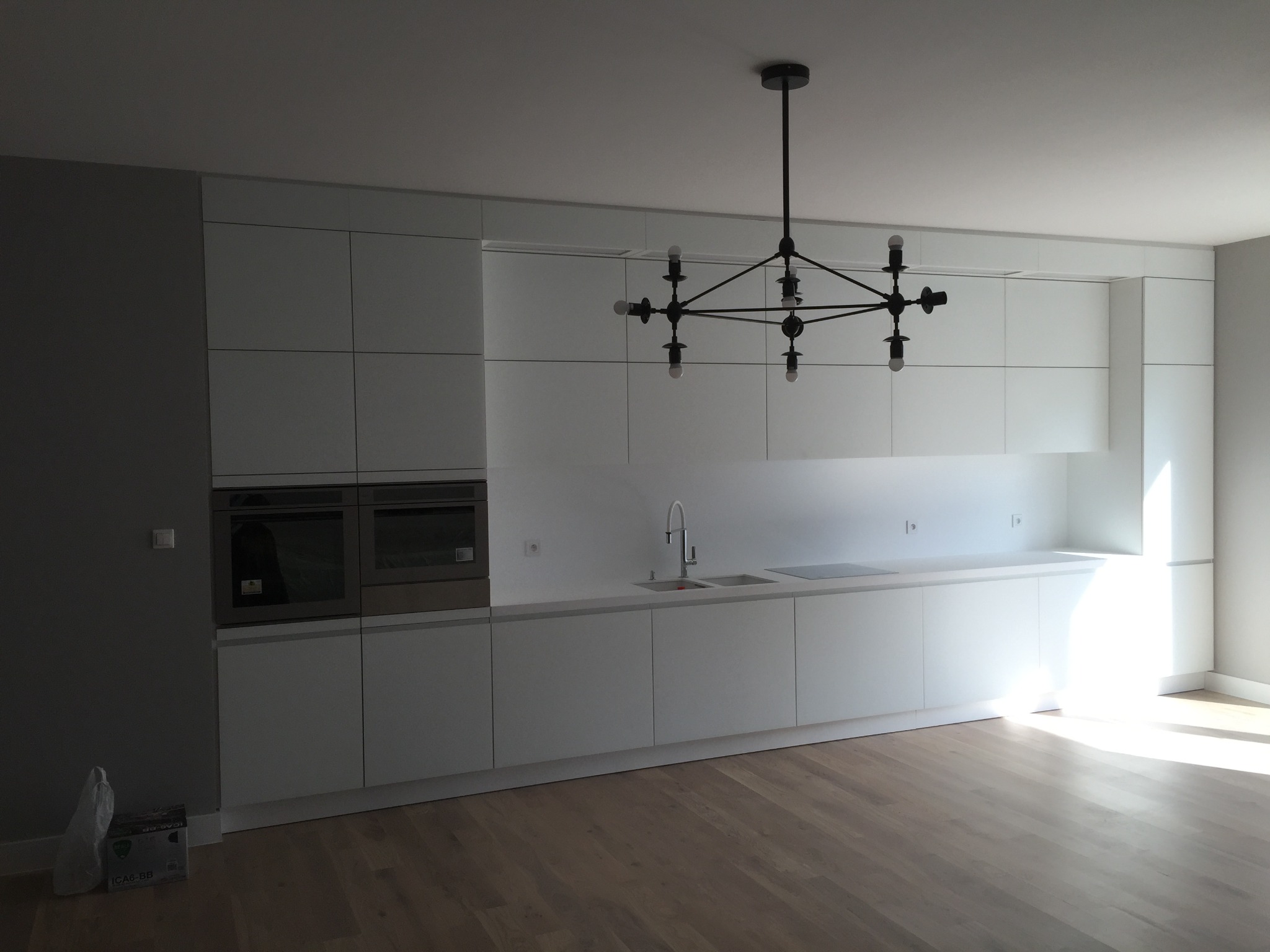 Minimalistyczna kuchnia z białymi, matowymi frontami szafek, drewnianą podłogą i czarnym, nowoczesnym żyrandolem; widoczne wbudowane piekarniki i zlewozmywak z baterią.