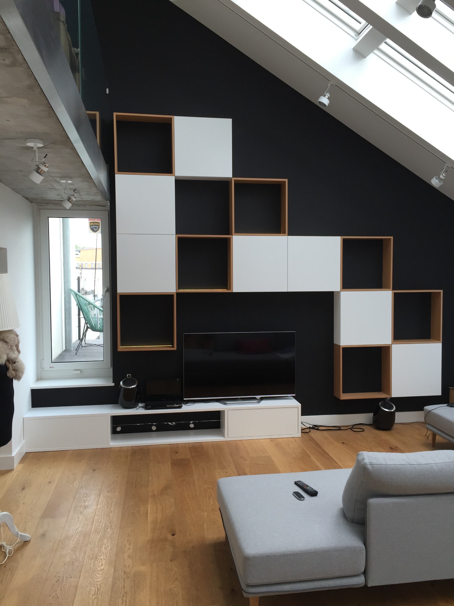 Nowoczesny salon na poddaszu z czarną ścianą, drewnianą podłogą i biało-drewnianą zabudową meblową w kształcie schodów, podświetlany sufit z oknami dachowymi i widoczne elementy szarej sofy