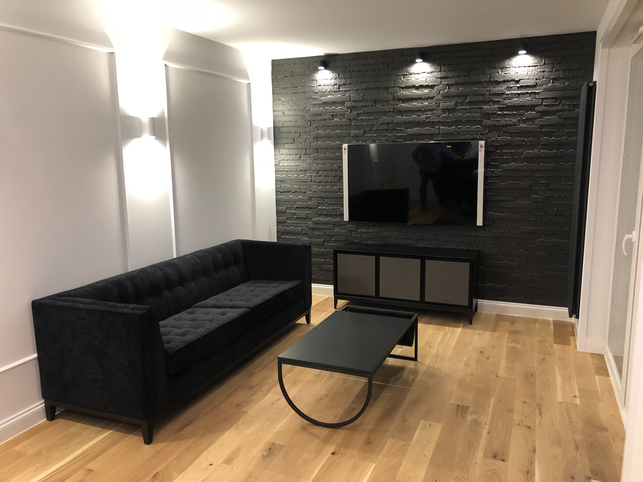 Salon z czarną sofą, stolikiem kawowym z metalową podstawą, szafką RTV i ścianą z czarnej cegły dekoracyjnej, oświetlony punktowymi lampami.