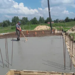 Wylewanie betonu na przygotowane zbrojenie fundamentu domu jednorodzinnego, pracownik wyrównuje powierzchnię za pomocą łaty, w tle widoczny betonowóz i sterta piasku.